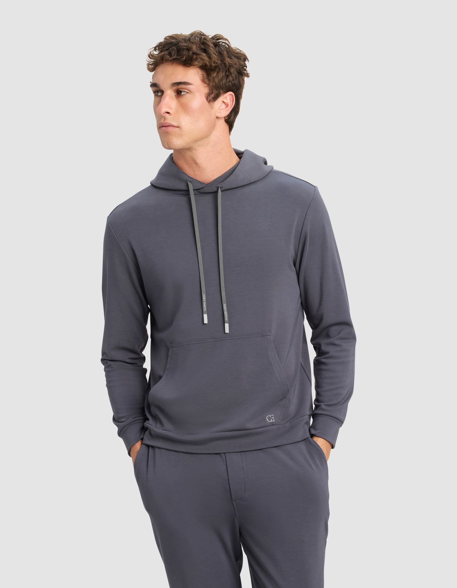 A young man models the Cozy Earth Men's Brushed Bamboo Hoodie in gray, standing with his hands in his pockets against a plain light background and looking to his left.|Color:Graphite