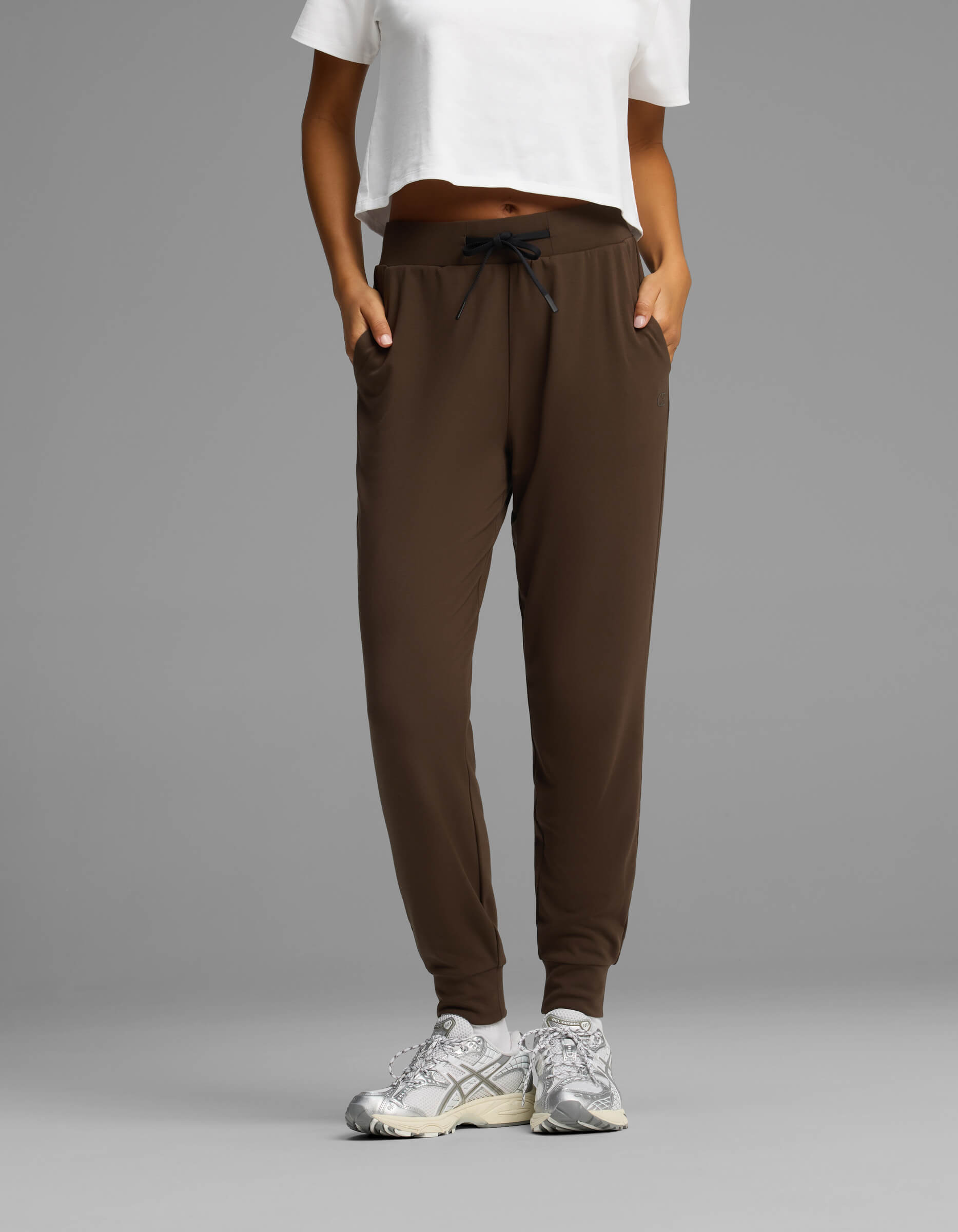 A person wearing Cozy Earth Women's Brushed Bamboo Joggers in brown, paired with a white cropped T-shirt and white sneakers, stands with hands in pockets against a plain gray background.