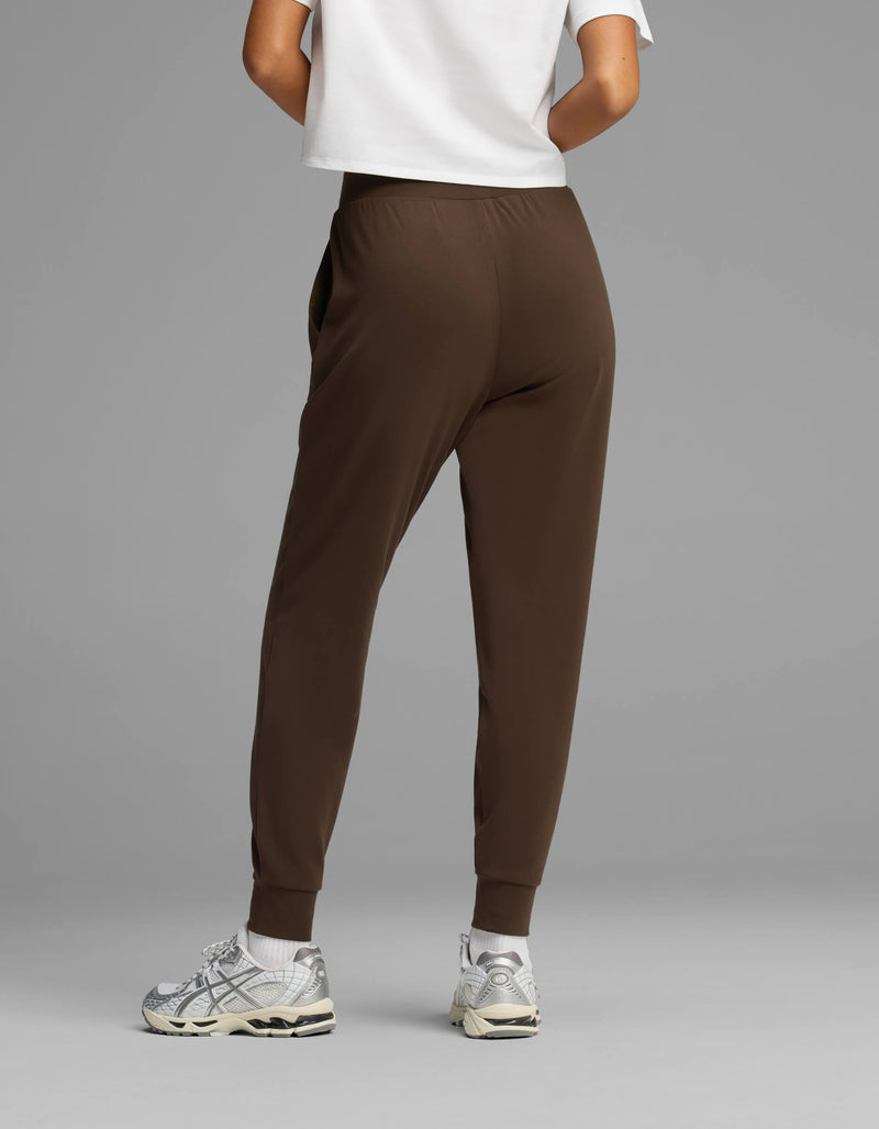 A person stands facing away against a plain gray background, wearing Cozy Earth Women's Brushed Bamboo Joggers in brown, paired with a white shirt and white sneakers.