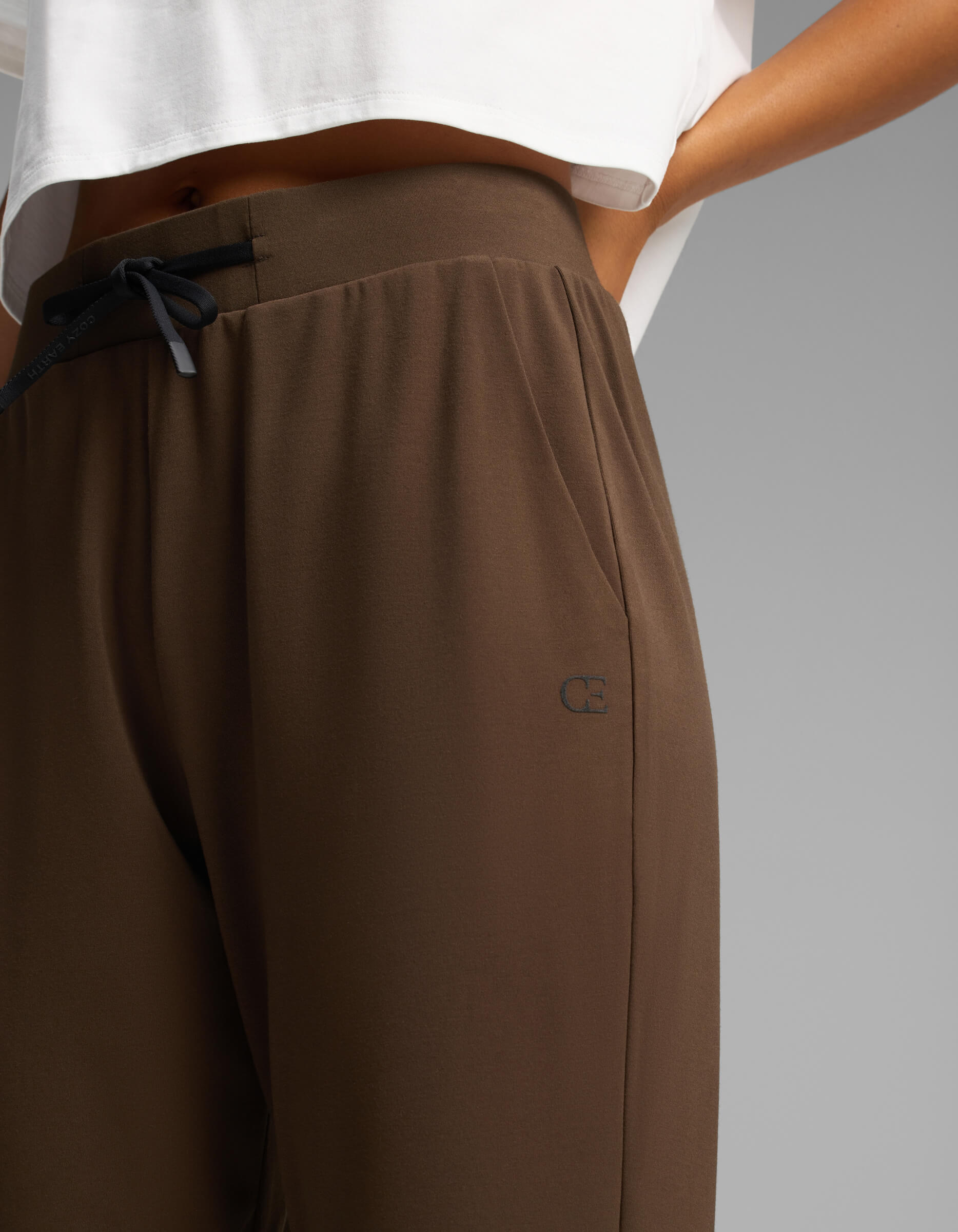 A person stands with one hand in their pocket, wearing Cozy Earth Women's Brushed Bamboo Joggers in brown paired with a white cropped shirt. The joggers feature a small logo near the pocket.