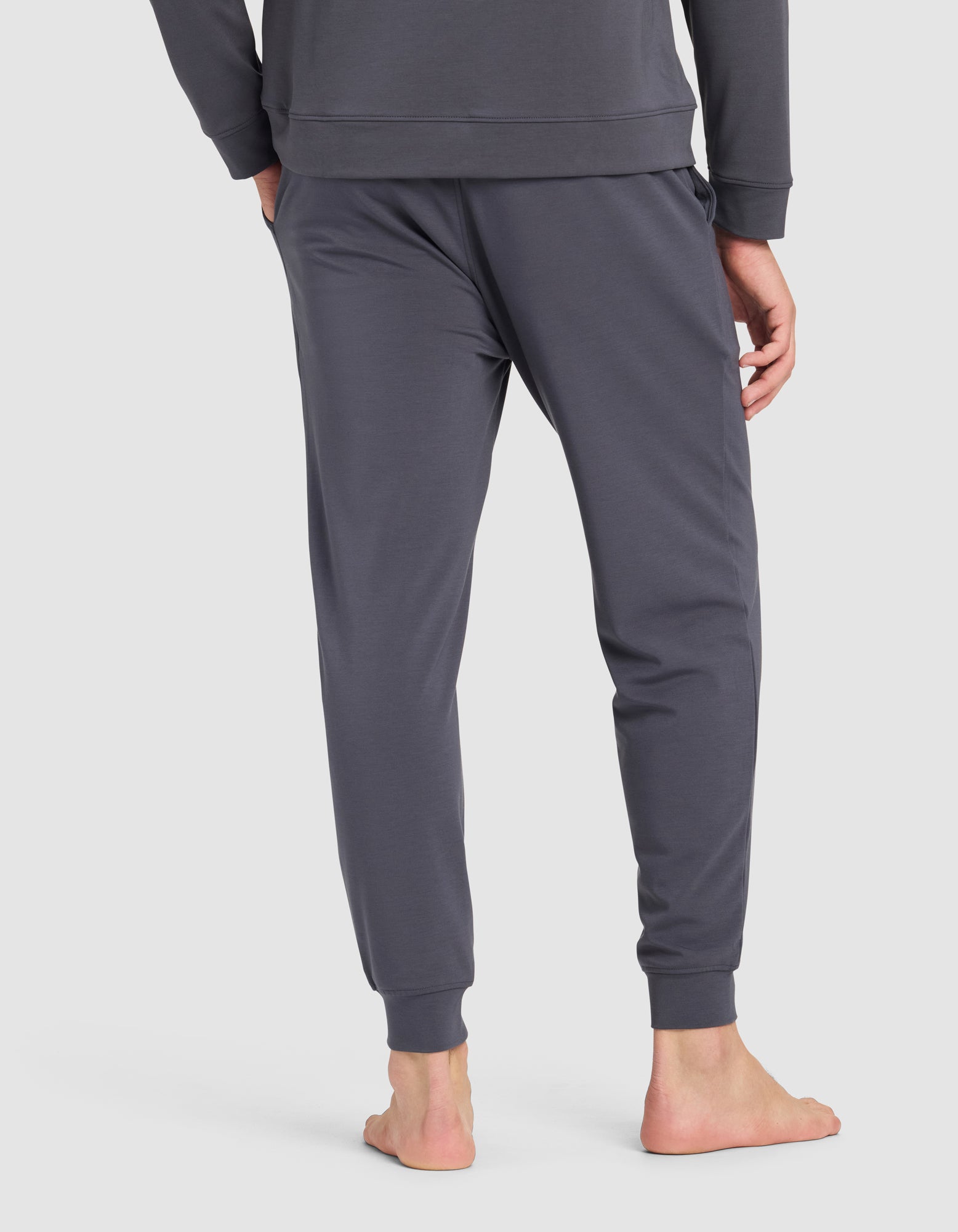 A man stands barefoot, seen from behind, wearing Cozy Earth Men's Brushed Bamboo Jogger Pant in dark gray, paired with a matching long-sleeve top, against a plain light background.