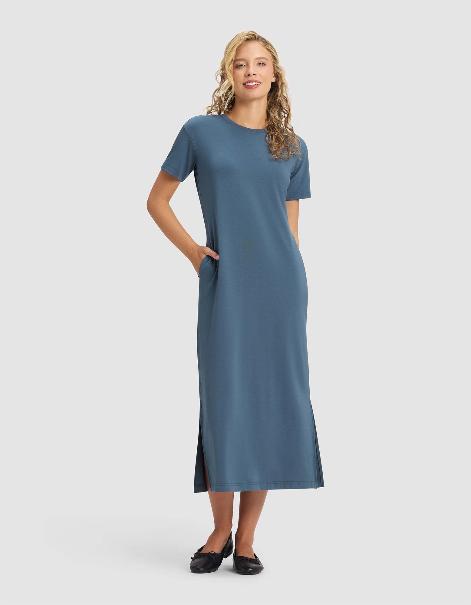 A woman smiles in the Cozy Earth Women's Brushed Bamboo Midi Dress, a blue, short-sleeve, ankle-length style with side slits and pockets. She pairs it with black flats. Her long blonde hair is loose against a plain light gray background. 
