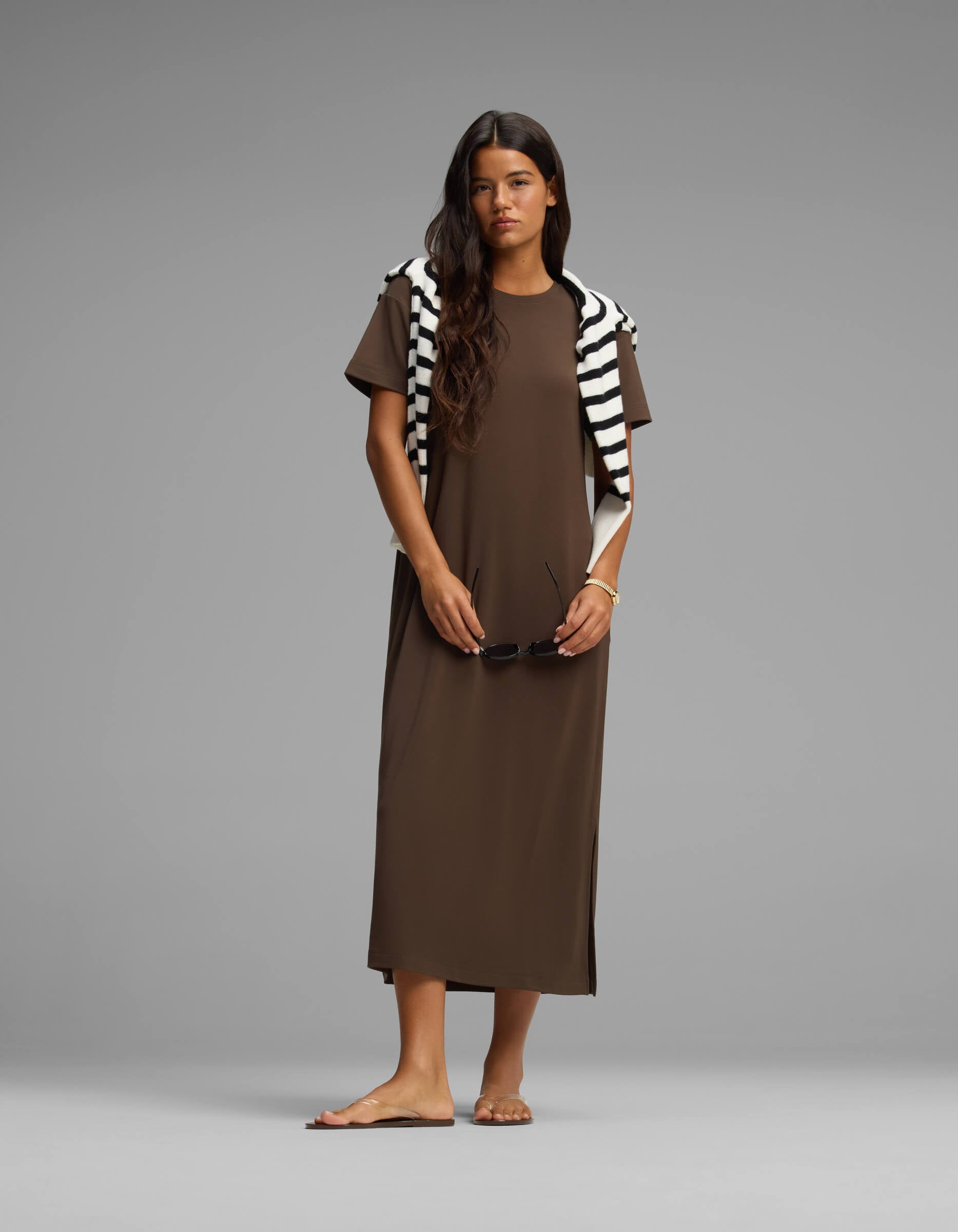 A woman stands barefoot against a plain gray background, wearing the Cozy Earth Women's Brushed Bamboo Midi Dress in brown with a black and white striped sweater over her shoulders, holding sunglasses in one hand.|Color:Espresso