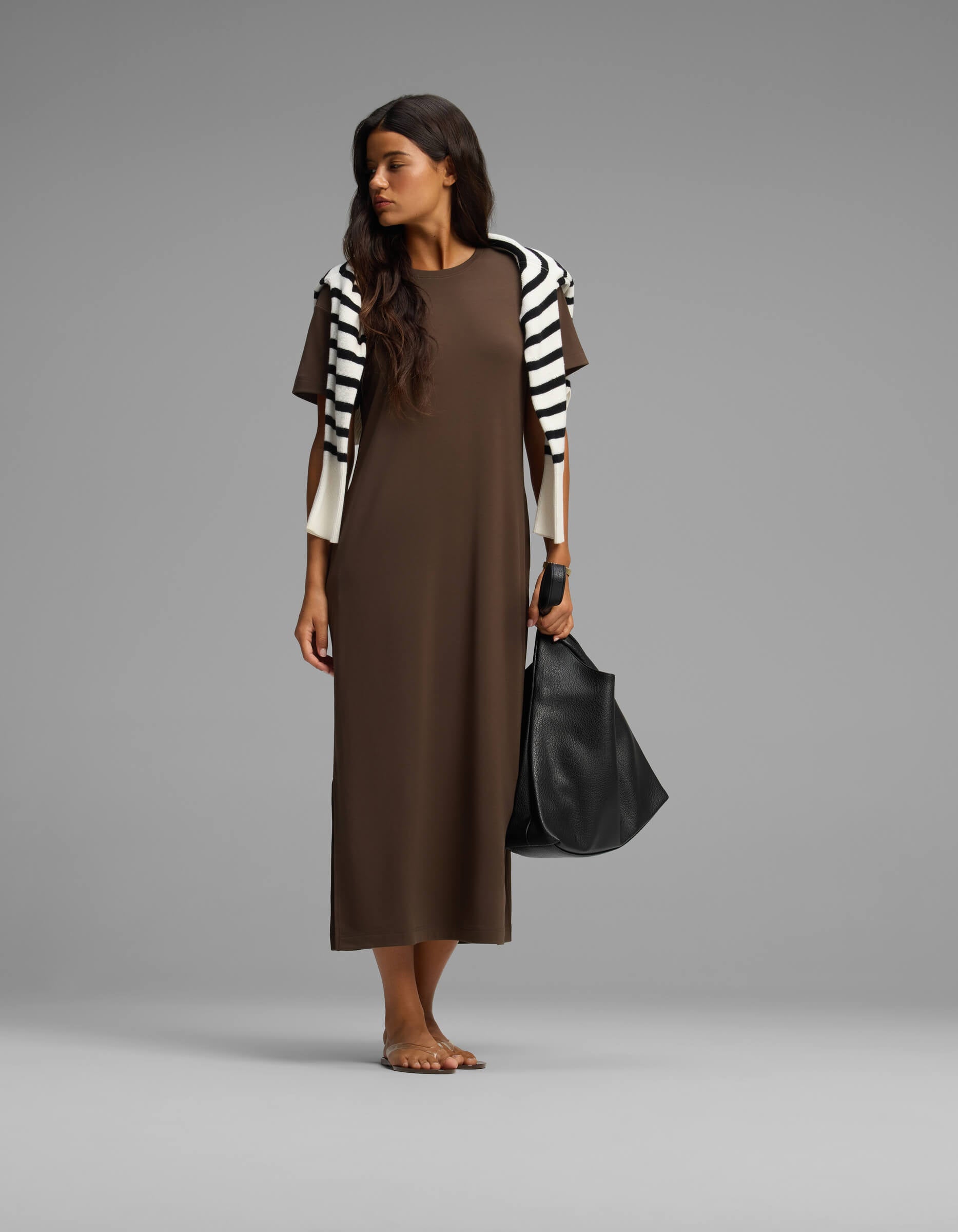A woman stands barefoot against a plain background, wearing Cozy Earth’s Women's Brushed Bamboo Midi Dress in brown, with a black-and-white striped sweater over her shoulders and holding a large black bag.