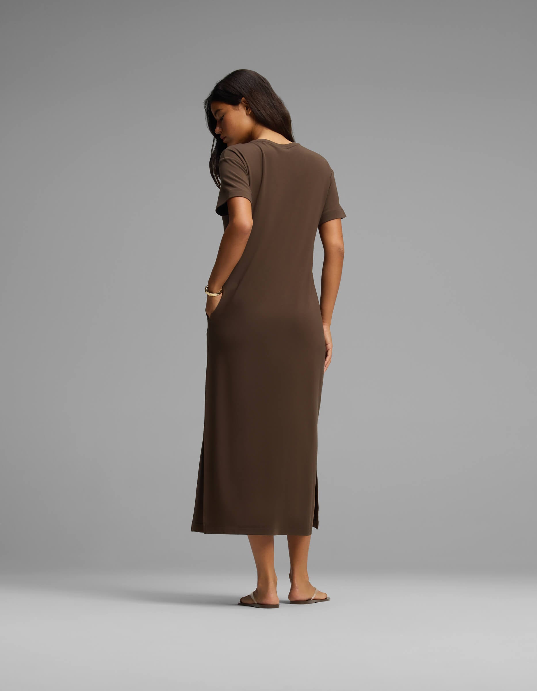 A woman stands barefoot facing away, wearing Cozy Earth's Women's Brushed Bamboo Midi Dress—a short-sleeve, ankle-length brown dress with her right hand in her pocket—set against a plain light gray background.