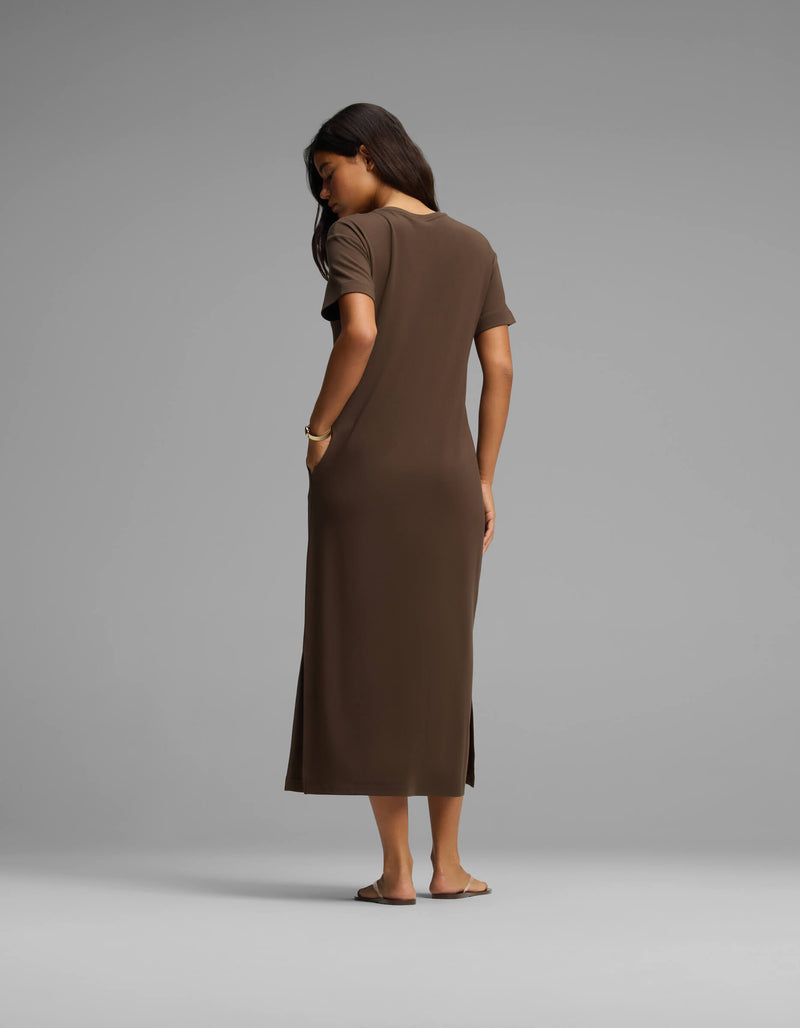 A woman stands barefoot facing away, wearing Cozy Earth's Women's Brushed Bamboo Midi Dress—a short-sleeve, ankle-length brown dress with her right hand in her pocket—set against a plain light gray background.