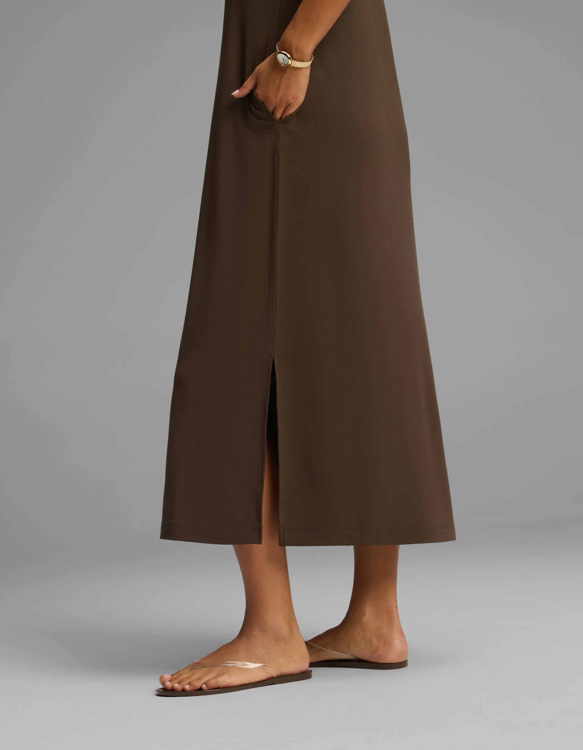 The lower half of a person is shown wearing the Cozy Earth Women's Brushed Bamboo Midi Dress in brown, featuring a center slit, paired with a bracelet and clear flat sandals, standing against a plain gray background.