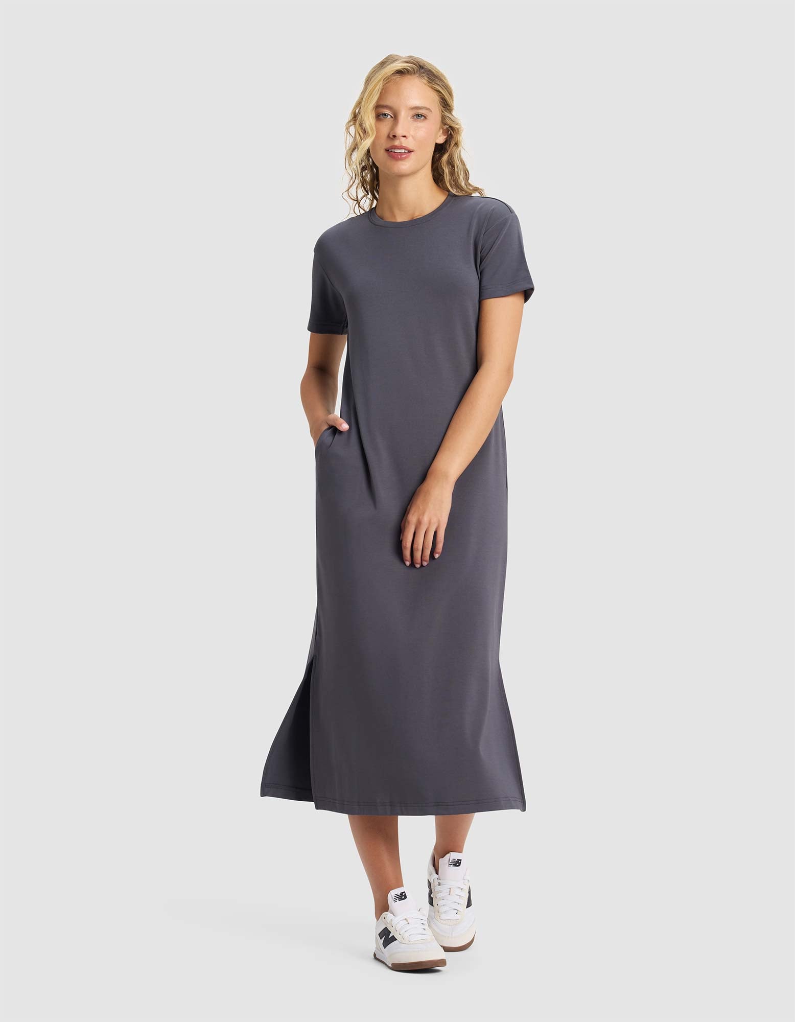 A woman smiles against a plain background, wearing the Cozy Earth Women's Brushed Bamboo Midi Dress in gray with short sleeves, side pockets, and white sneakers. She has one hand in her pocket.|Color:Graphite