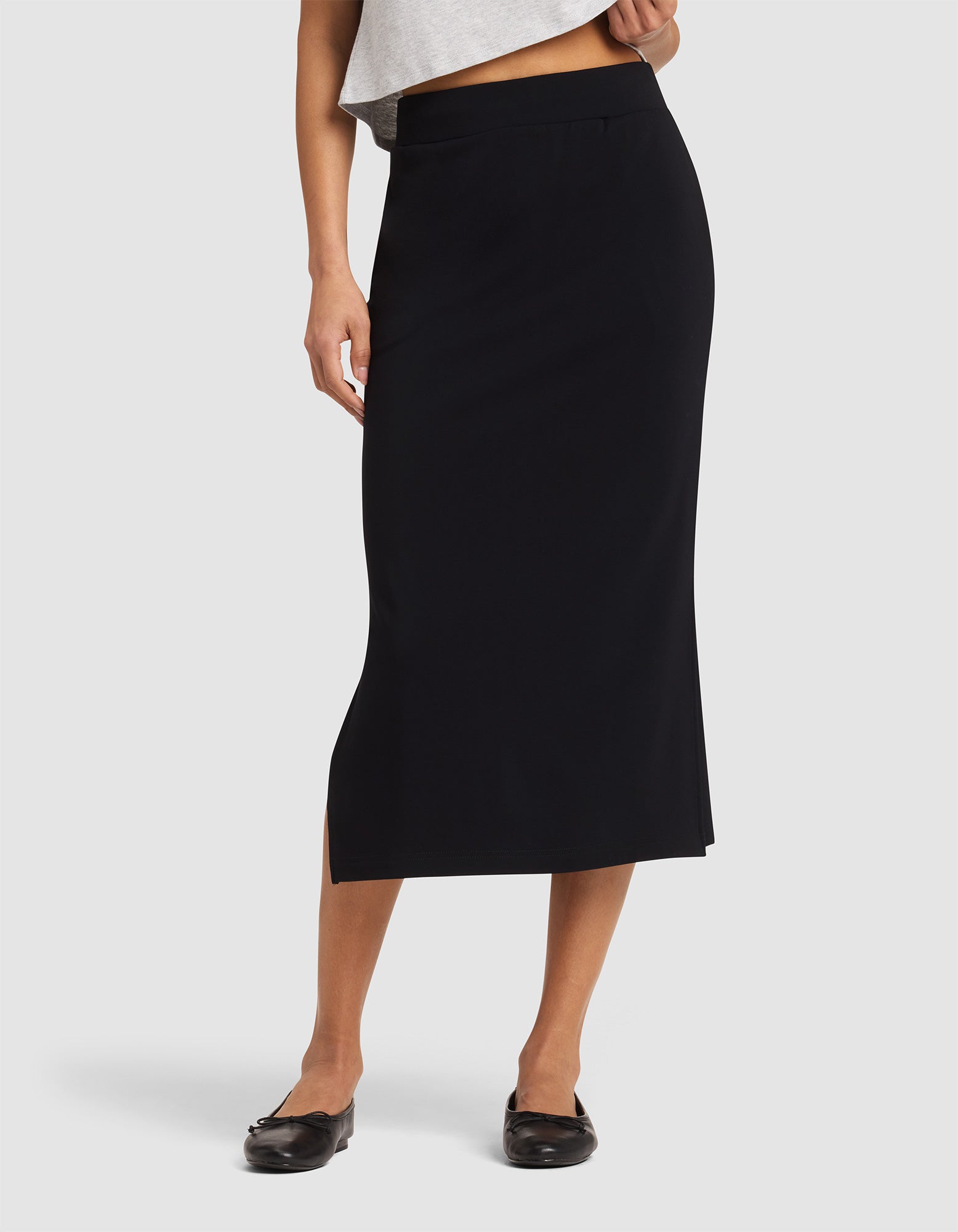 A woman wears the Cozy Earth Women's Brushed Bamboo Midi Skirt in black, high-waisted and ankle-length with a subtle side slit, paired with a gray top and black flats against a plain white background. 