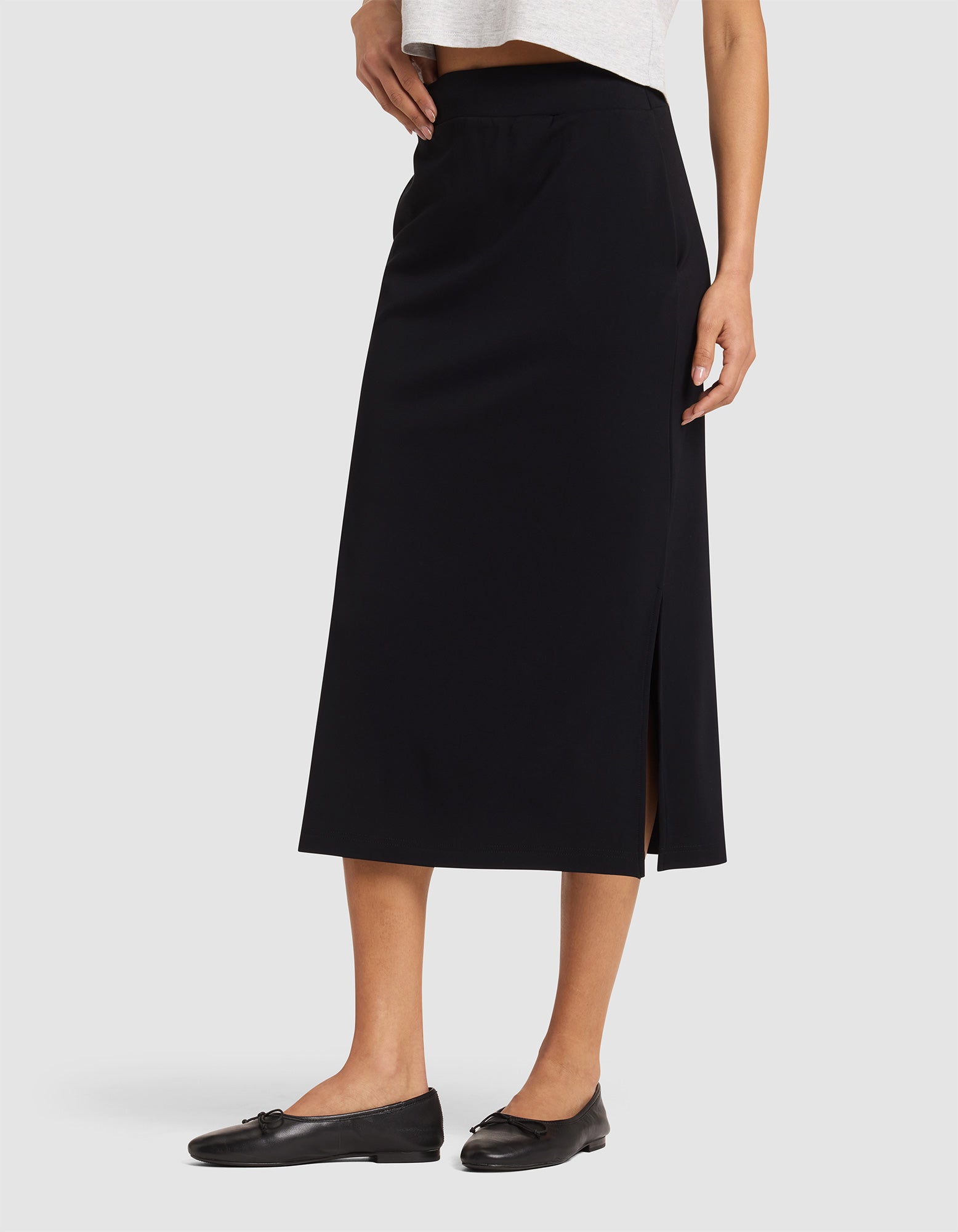 A woman wears the Cozy Earth Women's Brushed Bamboo Midi Skirt—black, knee-length with a side slit—paired with a white top and black flats against a plain light background. 