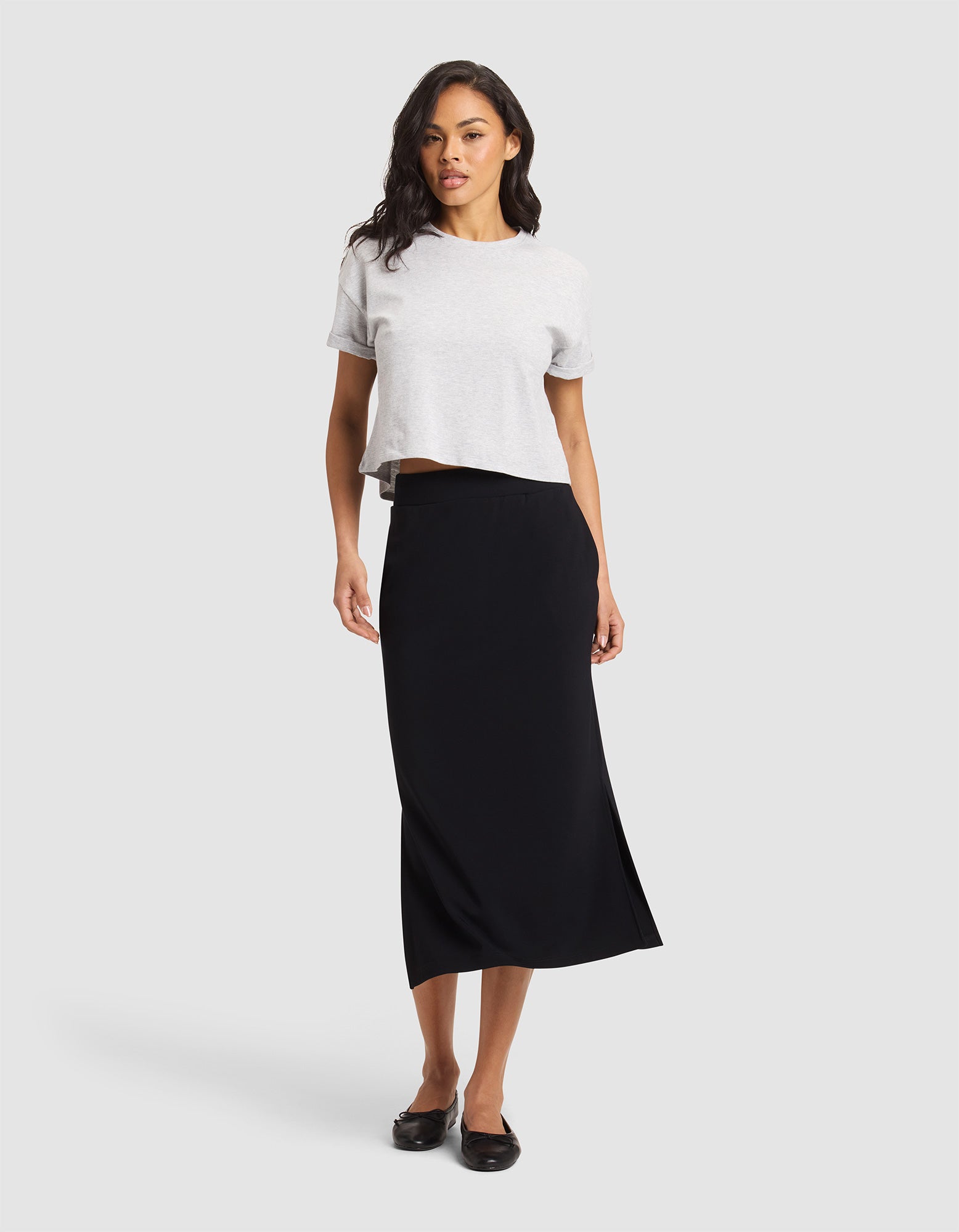 A woman with wavy dark hair and a confident stance wears the Cozy Earth Women's Brushed Bamboo Midi Skirt in black, paired with a light gray short-sleeve top and black flats, standing against a plain background. 