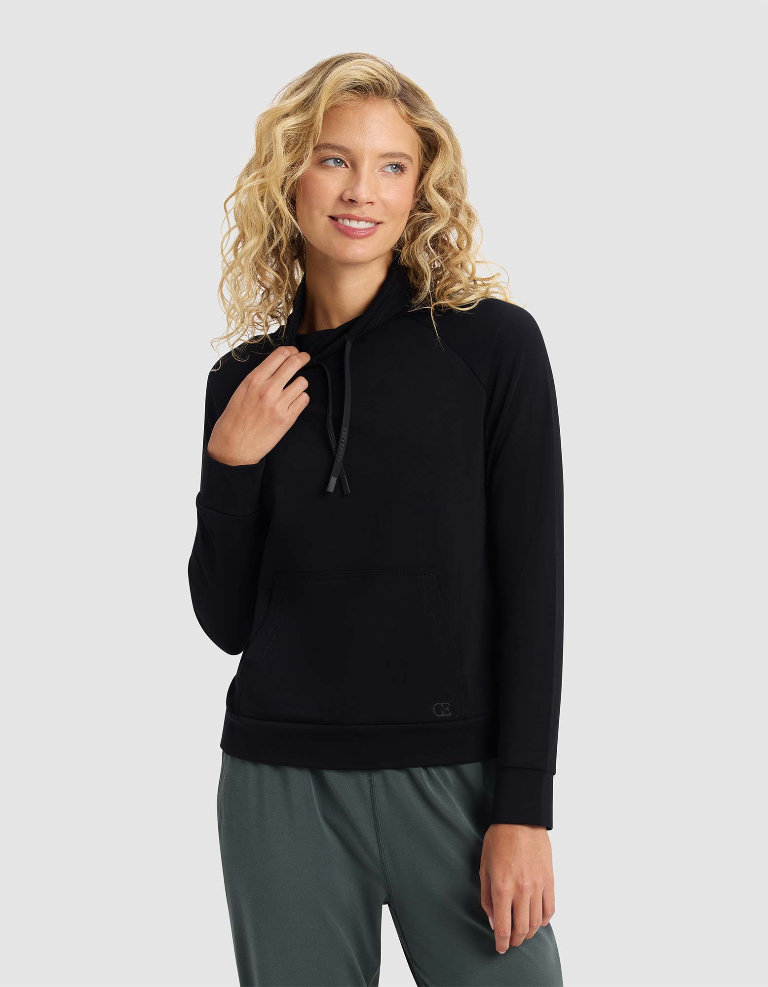 A smiling woman with blonde curly hair is wearing the Cozy Earth Women's Brushed Bamboo Mock Neck, paired with green pants, standing against a light gray background and looking to the side. 