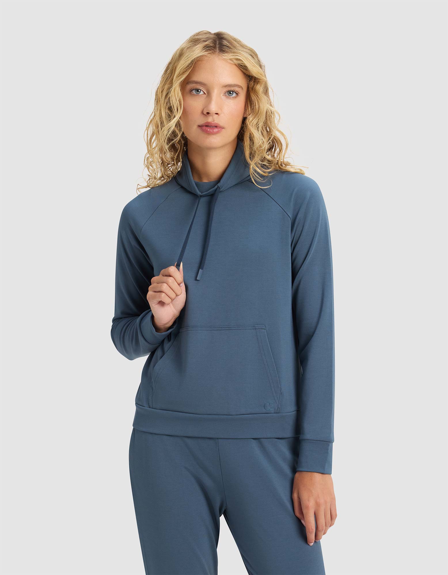 A woman with wavy blonde hair models the Cozy Earth Women's Brushed Bamboo Mock Neck in blue, standing against a plain light background. She holds the hoodie drawstring in one hand, wearing matching pants and a neutral expression.