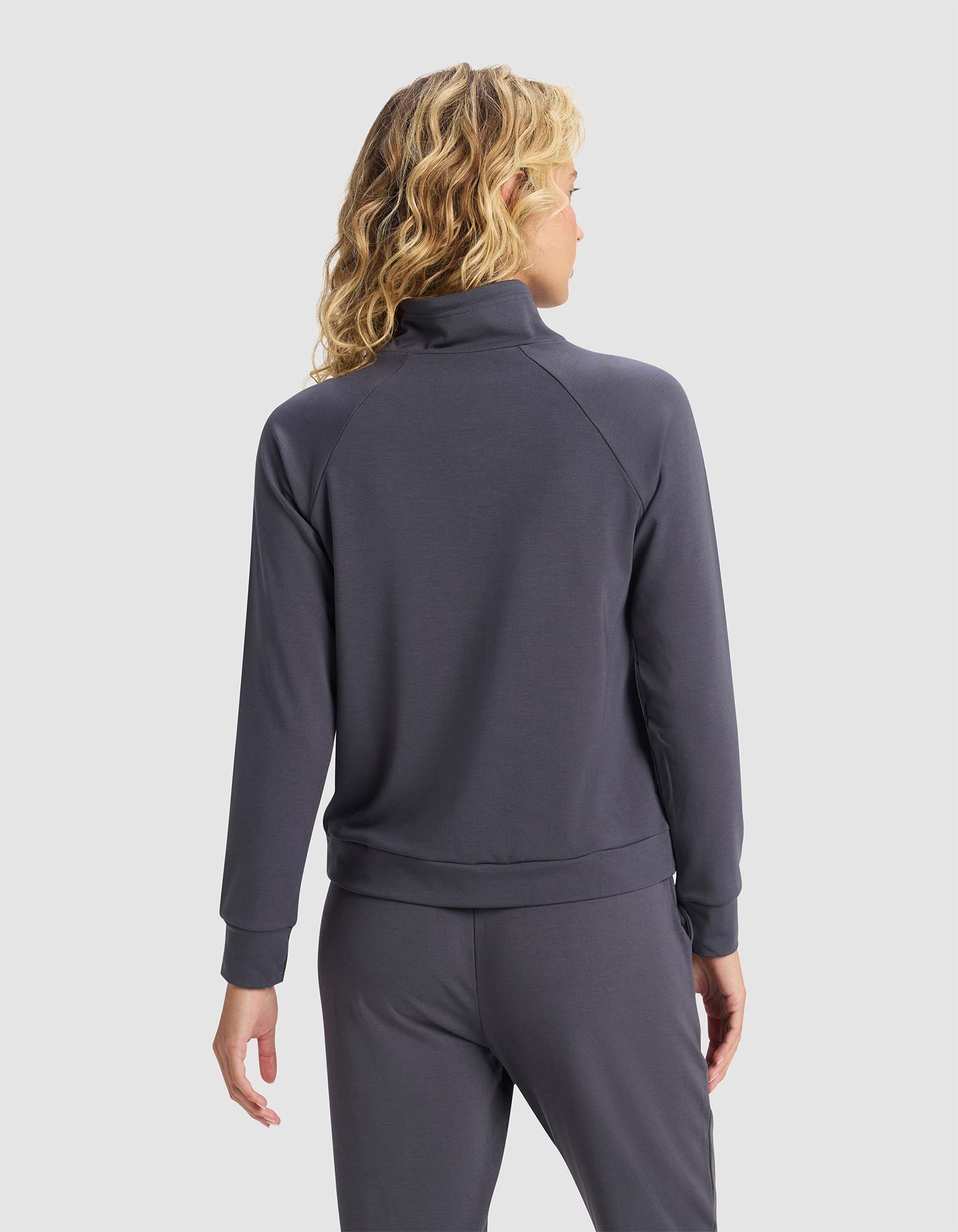 A person with wavy blonde hair models the back of Cozy Earth's Women's Brushed Bamboo Mock Neck in dark gray, along with matching pants, standing against a plain light background.|Color:Graphite