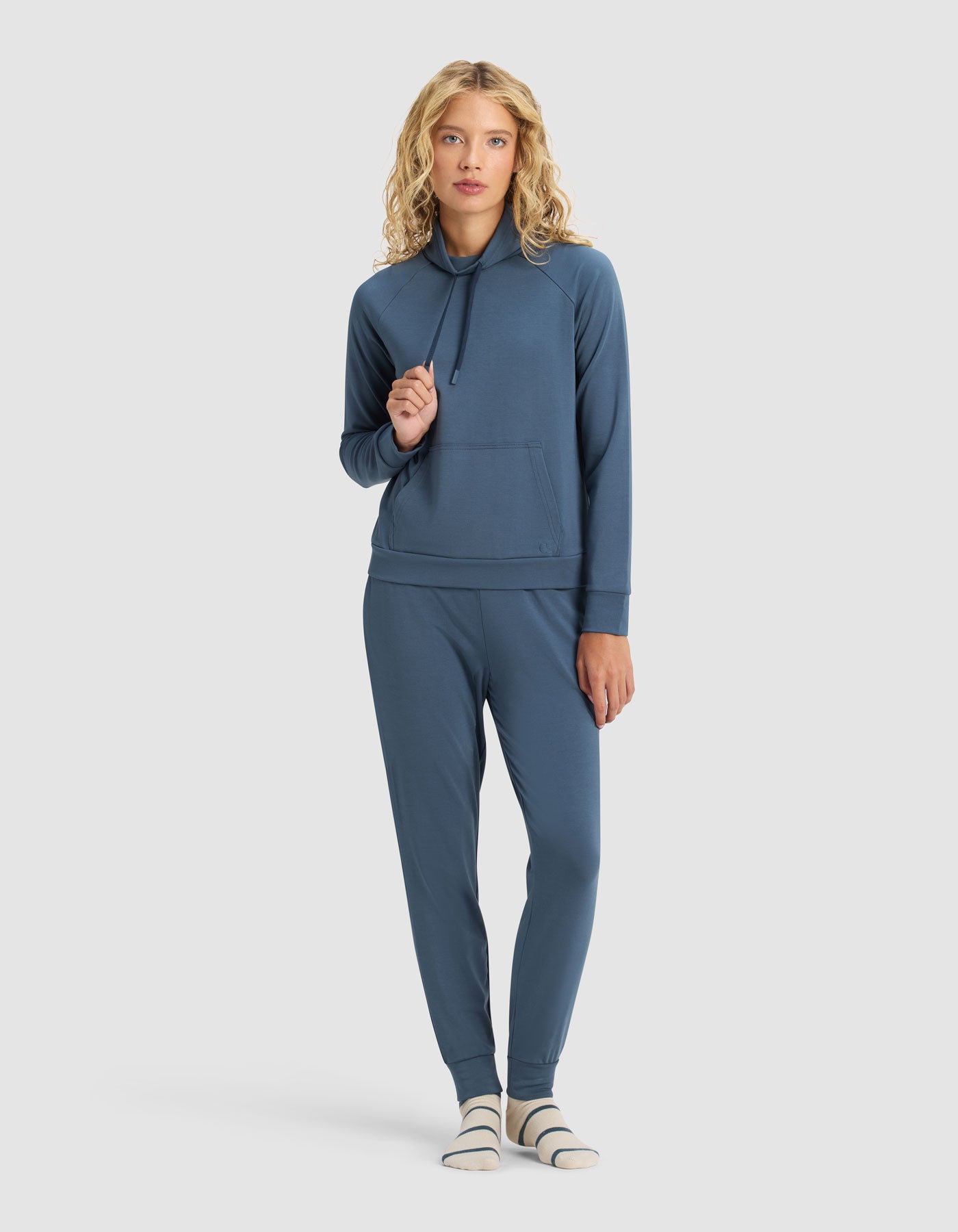 A woman with blonde curly hair wears the Cozy Earth Women's Brushed Bamboo Mock Neck & Jogger Set, paired with striped socks, standing against a plain light background.|Color:Dawn