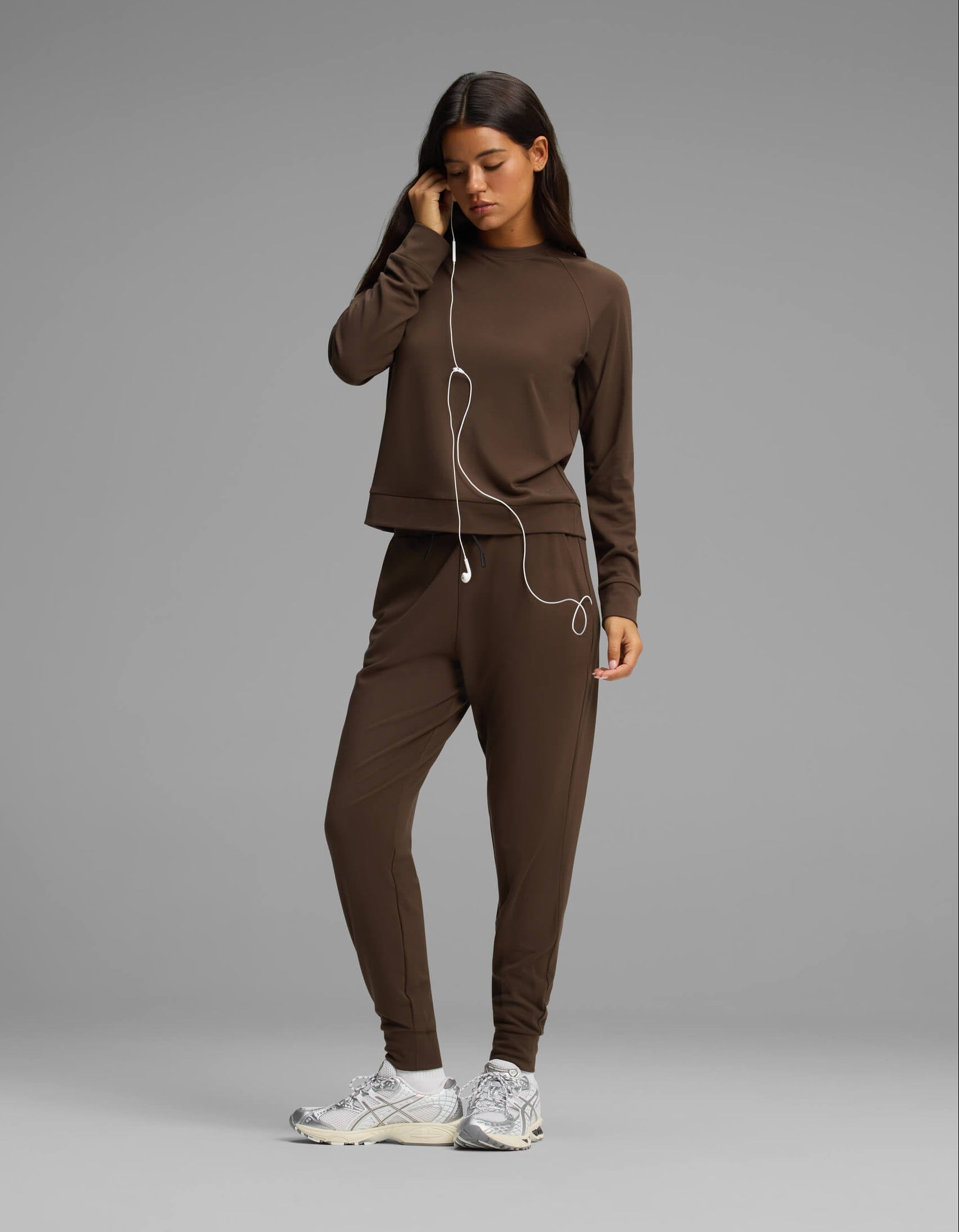 A woman wears the Cozy Earth Women's Brushed Bamboo Jogger Set, featuring a brown long-sleeve top and matching pants, as she stands in sneakers and holds white earphones to her ear against a plain gray background.|Color:Espresso