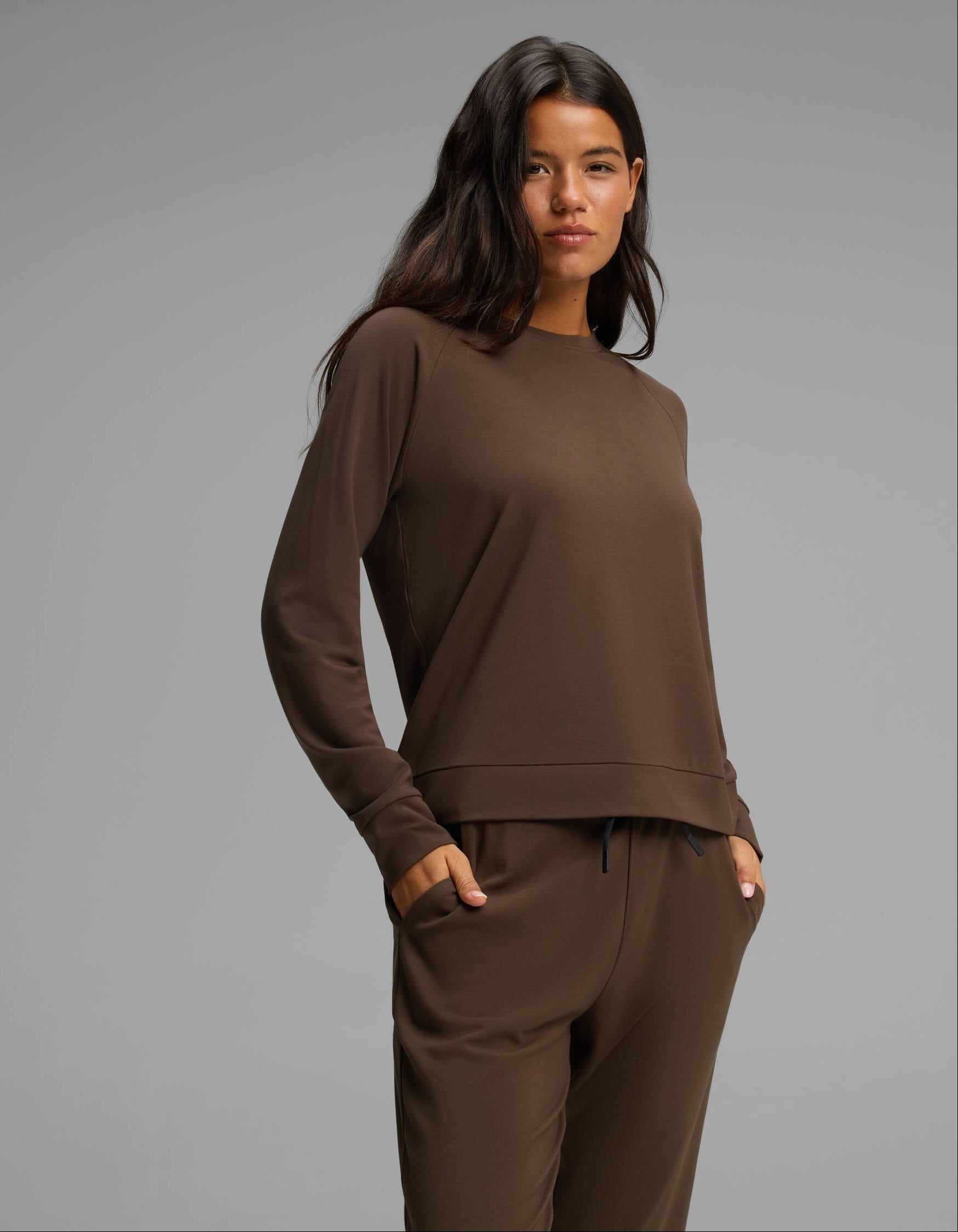 A woman with long dark hair stands confidently with one hand in her pocket against a plain light gray background, wearing the Cozy Earth Women's Brushed Bamboo Jogger Set in a matching brown shade.