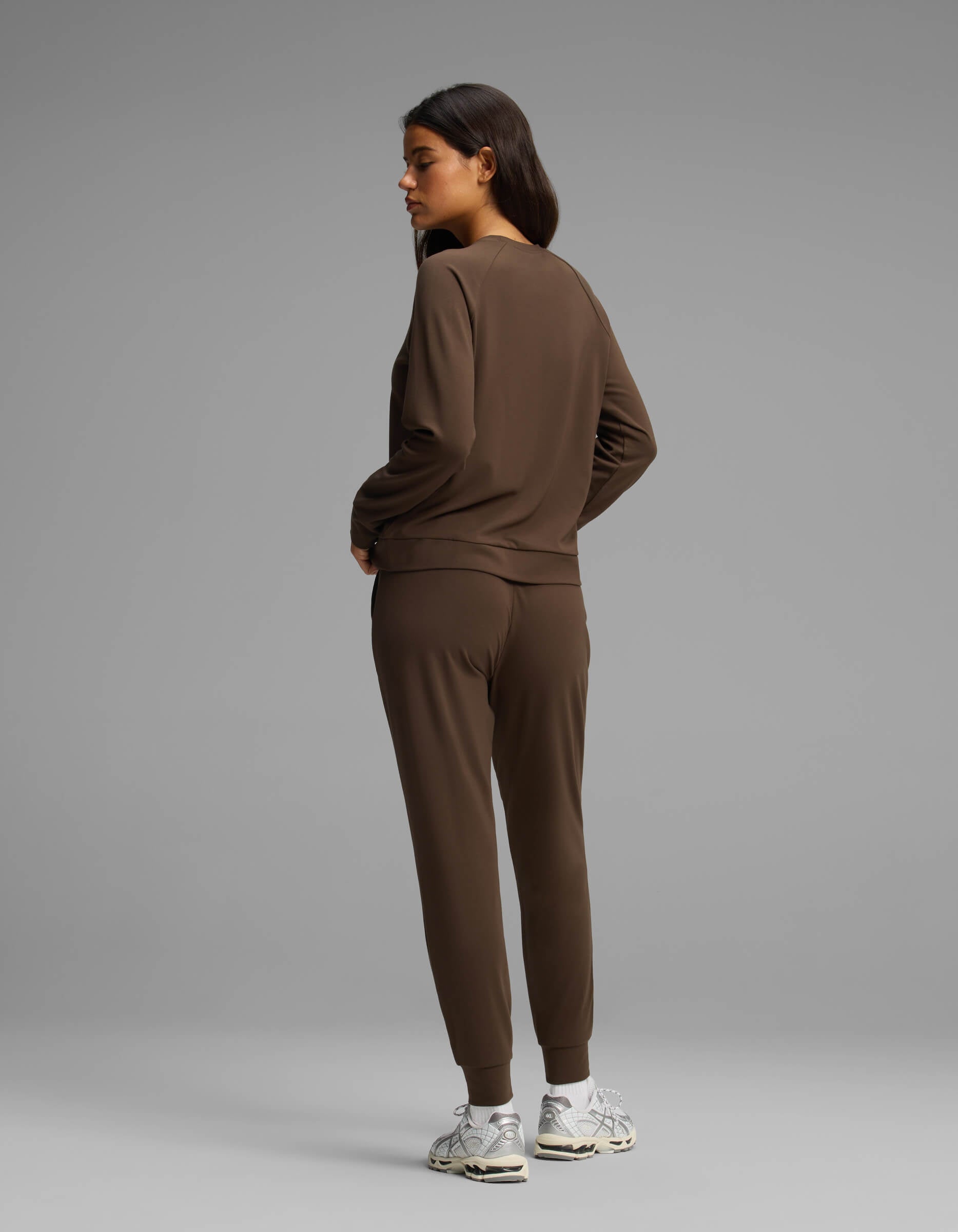 A woman models the Cozy Earth Women's Brushed Bamboo Jogger Set, featuring a brown long-sleeve top and matching jogger pants. She faces away and glances to her left, wearing white sneakers against a plain gray background.|Color:Espresso
