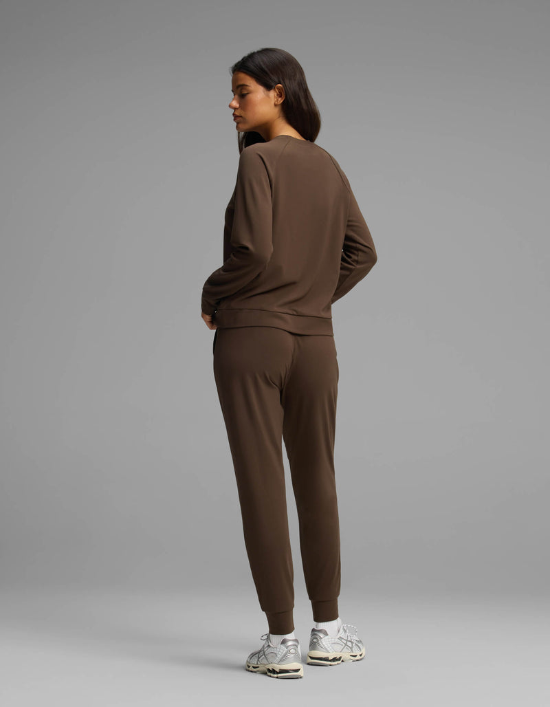 A woman models the Cozy Earth Women's Brushed Bamboo Jogger Set, featuring a brown long-sleeve top and matching jogger pants. She faces away and glances to her left, wearing white sneakers against a plain gray background.