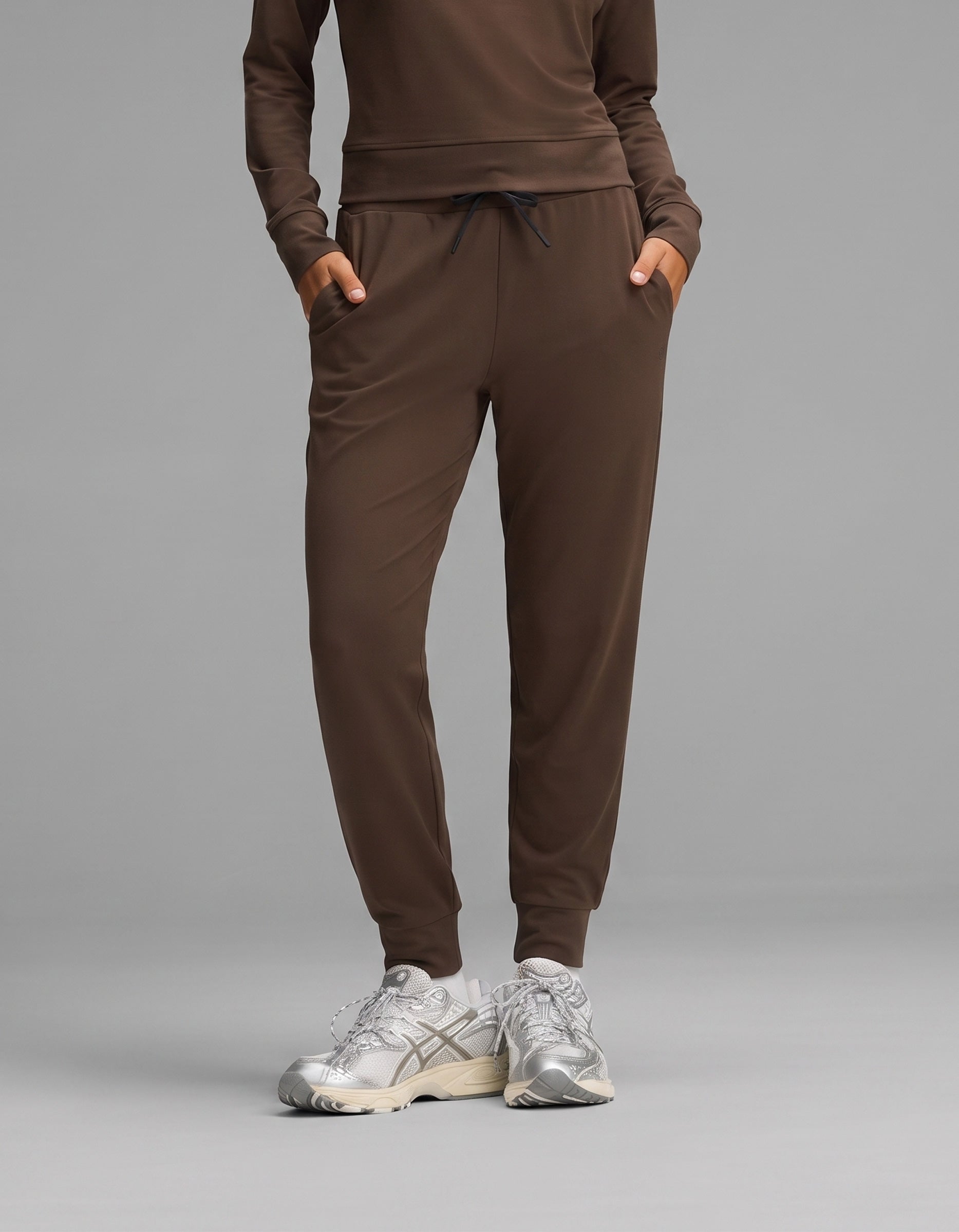 A person wearing the Cozy Earth Women's Brushed Bamboo Jogger Set in brown, paired with silver athletic sneakers, stands with hands in their pockets against a gray background.