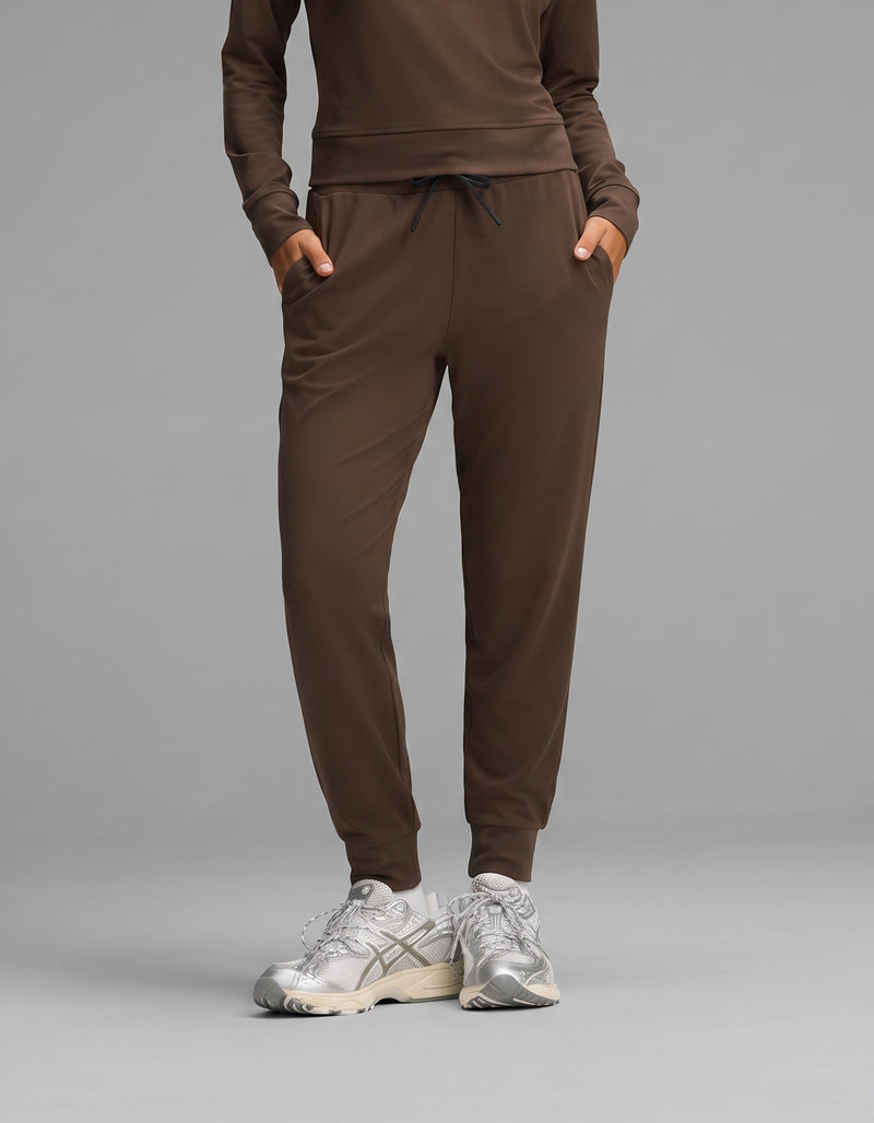 A person wearing the Cozy Earth Women's Brushed Bamboo Jogger Set in brown, paired with silver athletic sneakers, stands with hands in their pockets against a gray background.