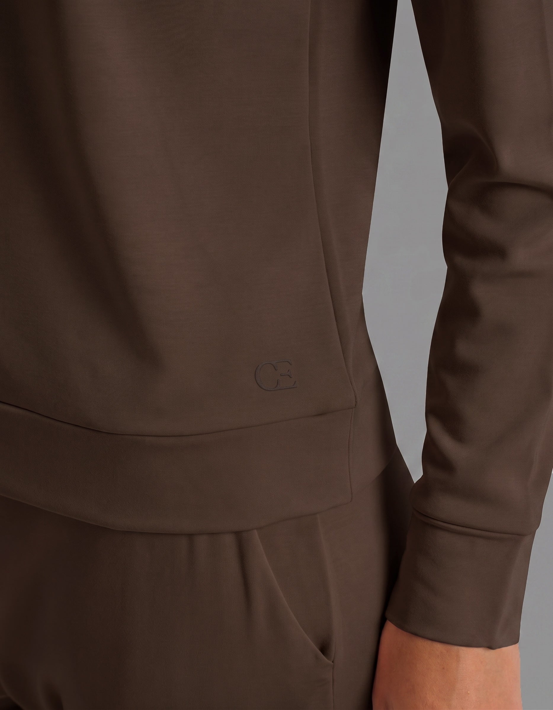 A person wearing the Cozy Earth Women's Brushed Bamboo Jogger Set in brown stands with one arm relaxed. The smooth fabric is complemented by a small embossed logo near the hem of the long-sleeve top.
