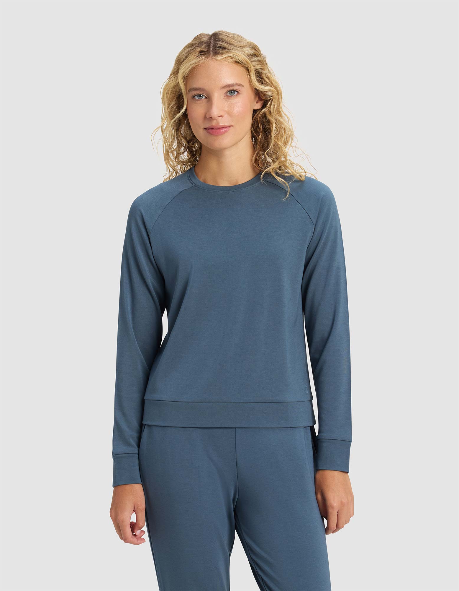 A woman with curly blonde hair faces forward against a plain light gray background, wearing the Cozy Earth Women's Brushed Bamboo Pullover Crew in blue, paired with matching pants.|Color:Dawn