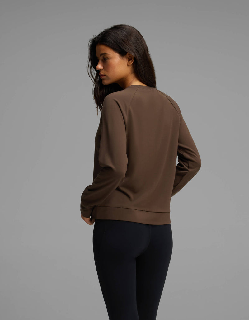 A woman with long dark hair, dressed in the Cozy Earth Women's Brushed Bamboo Pullover Crew and black leggings, stands turned slightly away from the camera against a plain gray background.