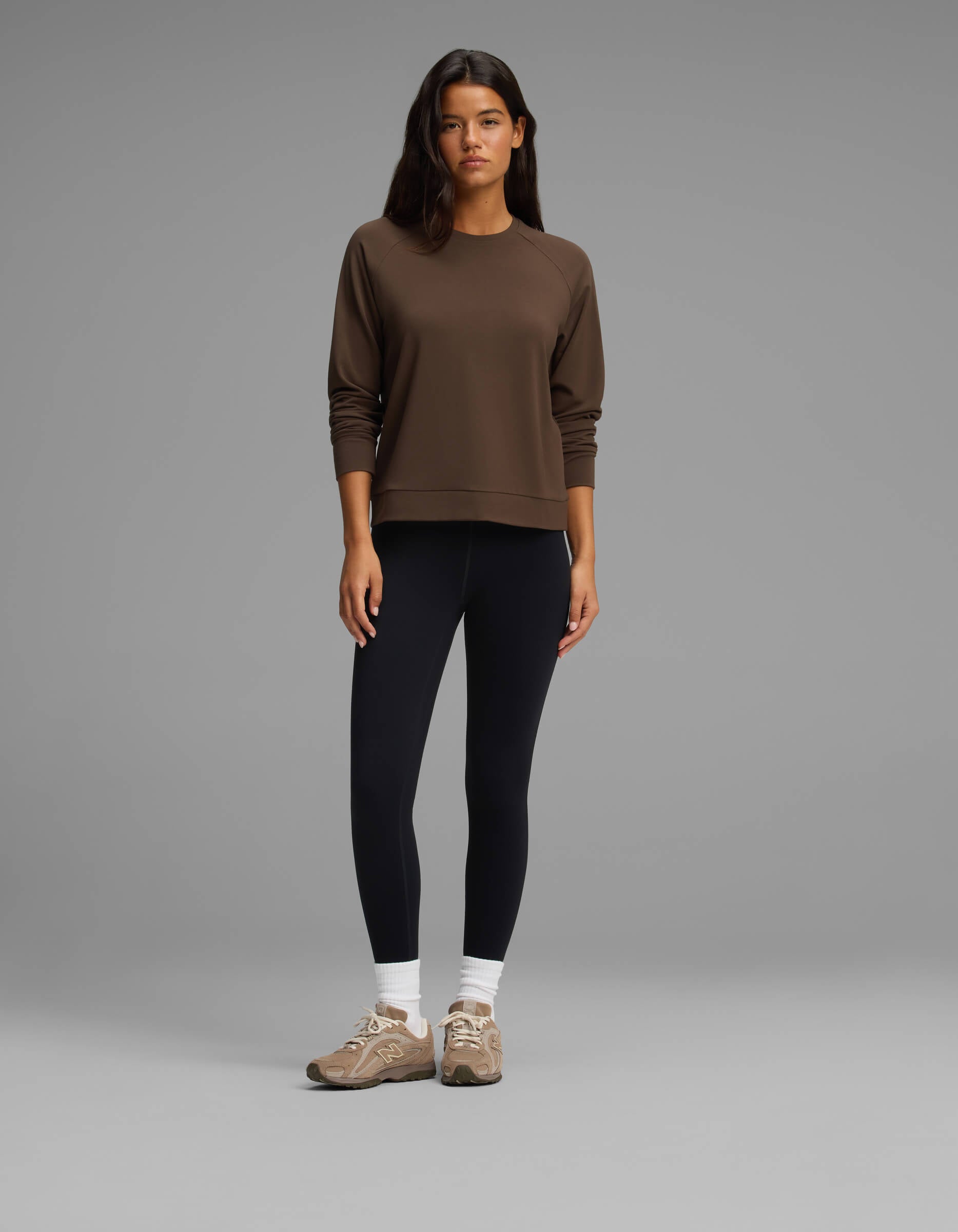 A woman stands against a gray background wearing the Cozy Earth Women's Brushed Bamboo Pullover Crew in brown, paired with black leggings, white socks, and tan sneakers. She looks at the camera with a neutral expression.