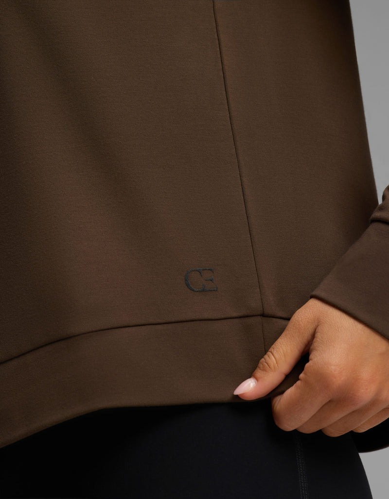 A person wearing the Cozy Earth Women's Brushed Bamboo Pullover Crew in brown gently pinches the hem with one hand, revealing a small, subtle embroidered "CB" logo near the bottom edge.