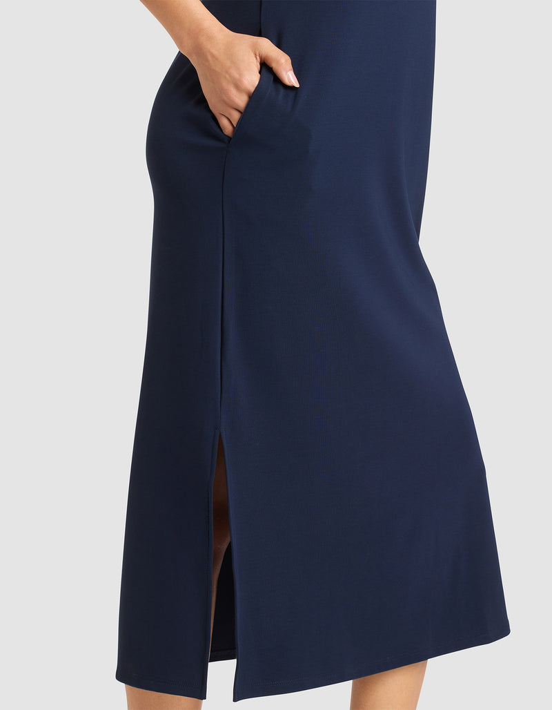 The lower half of a person is shown wearing the Cozy Earth Women's Brushed Bamboo Sleeveless Midi Dress in navy blue, featuring a side pocket and side slit, standing against a light gray background.