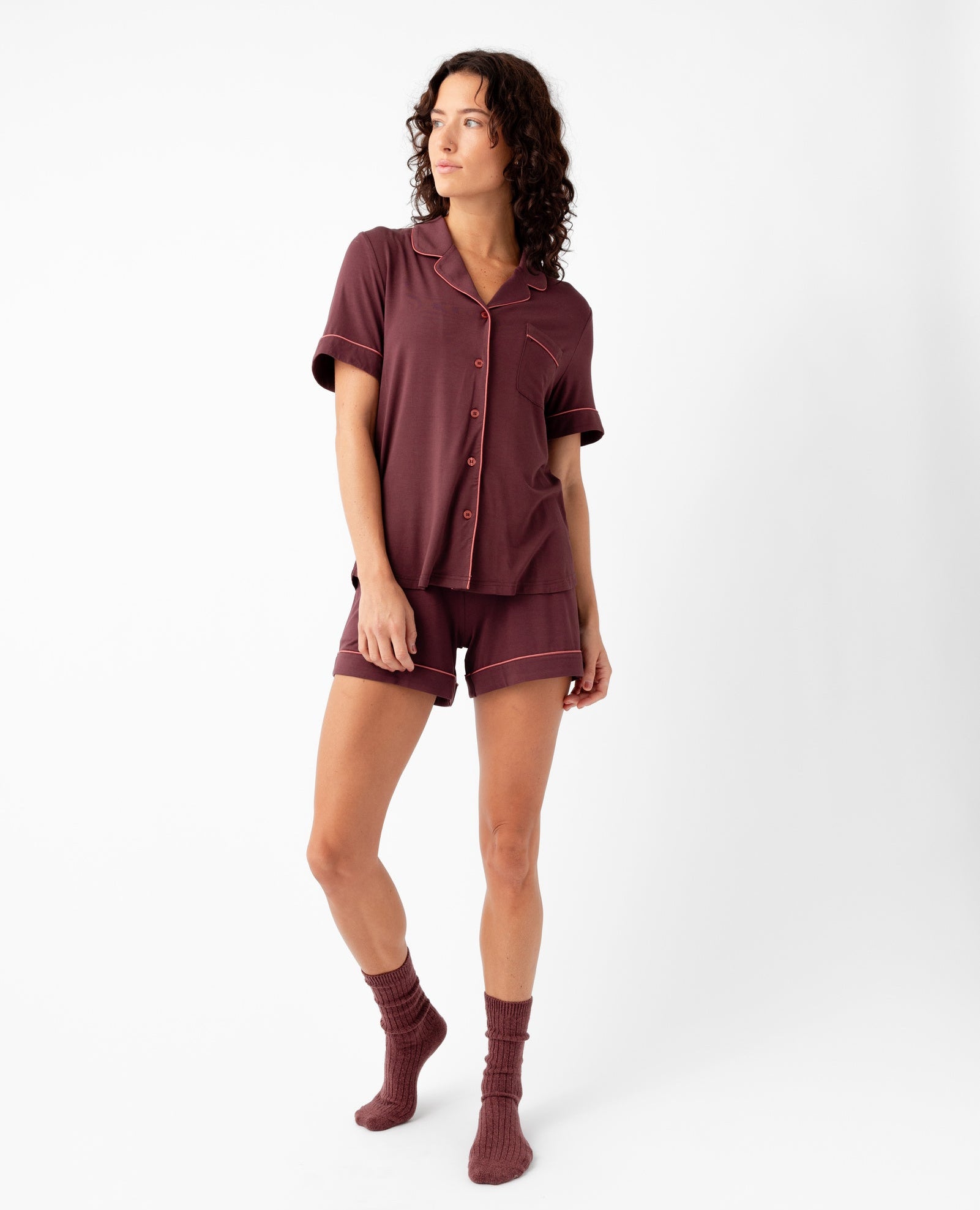 A woman with curly hair stands against a white background wearing Cozy Earth's Women's Bamboo Stretch-Knit Short Sleeve Pajama Set in burgundy with matching socks, looking to the side with a relaxed expression.