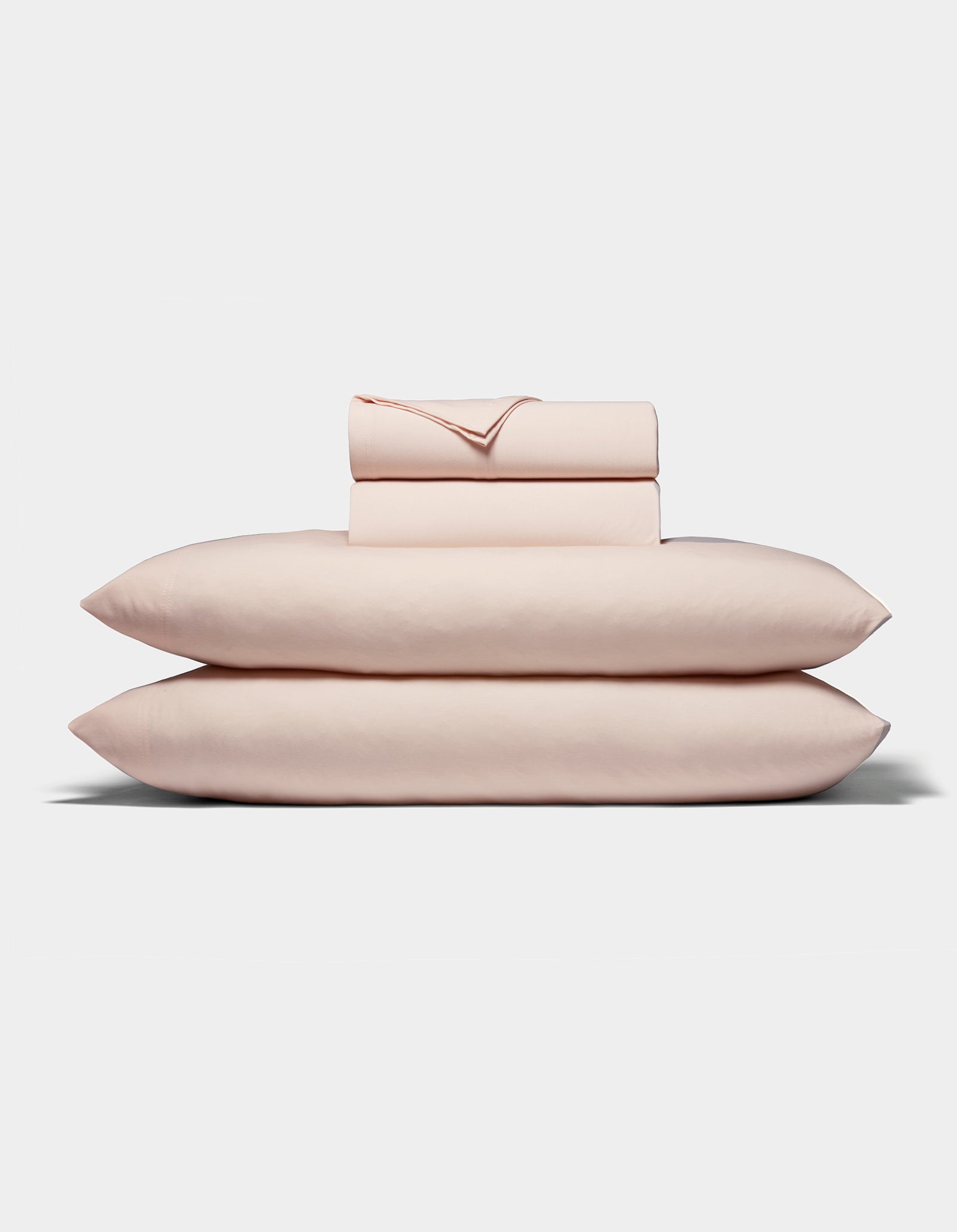 A neatly folded stack of Cozy Earth's Bamboo Jersey Sheet Set, in pale pink, featuring two pillows, a fitted sheet, a flat sheet, and pillowcases arranged on a white background.|Color:Petal
