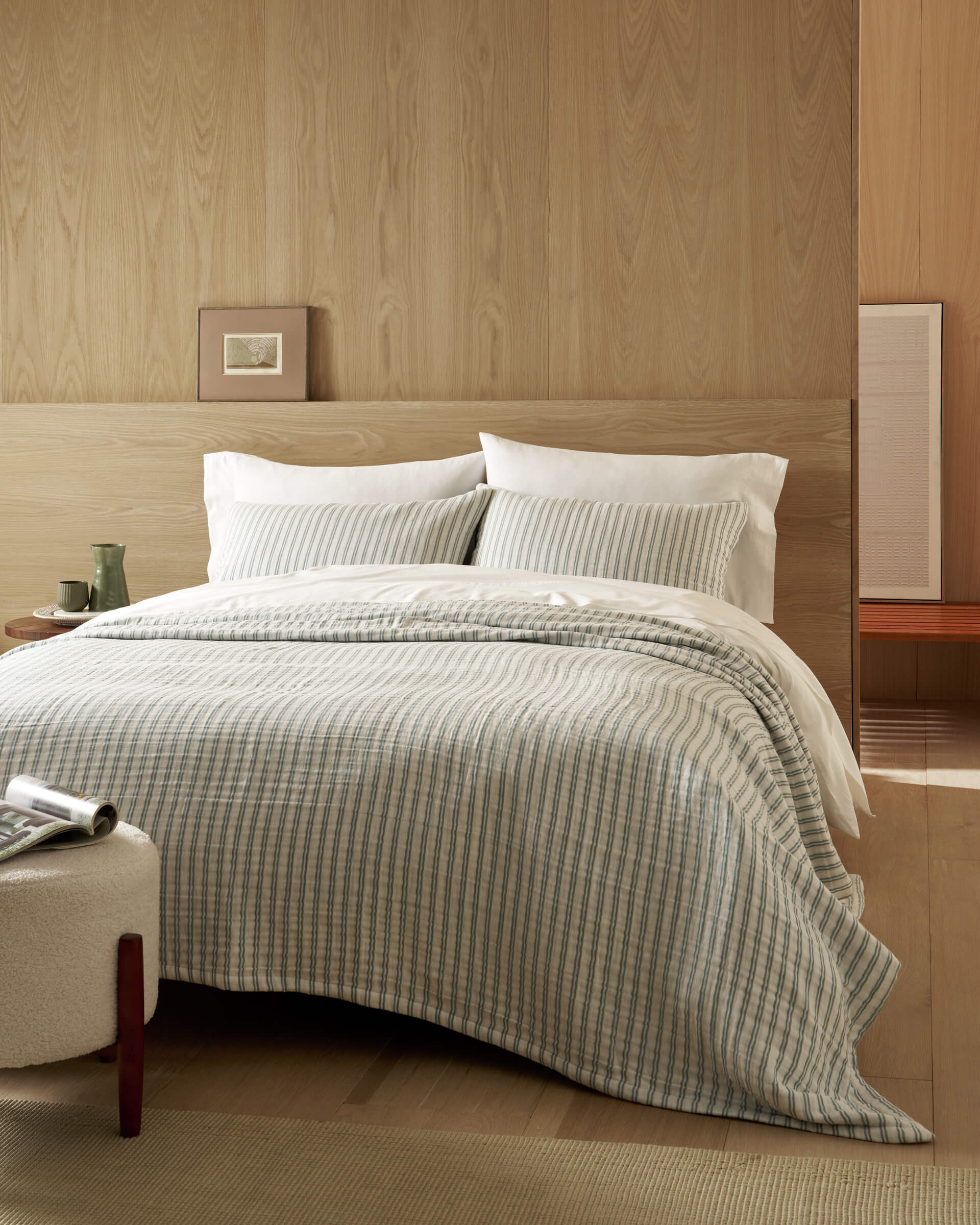A neatly made bed features the Cozy Earth Waterfront Coverlet against a light wood-paneled wall. White pillows, a small framed picture, a green cup on the table, and a bench complete this minimalist, cozy room.|Color:River Stripe