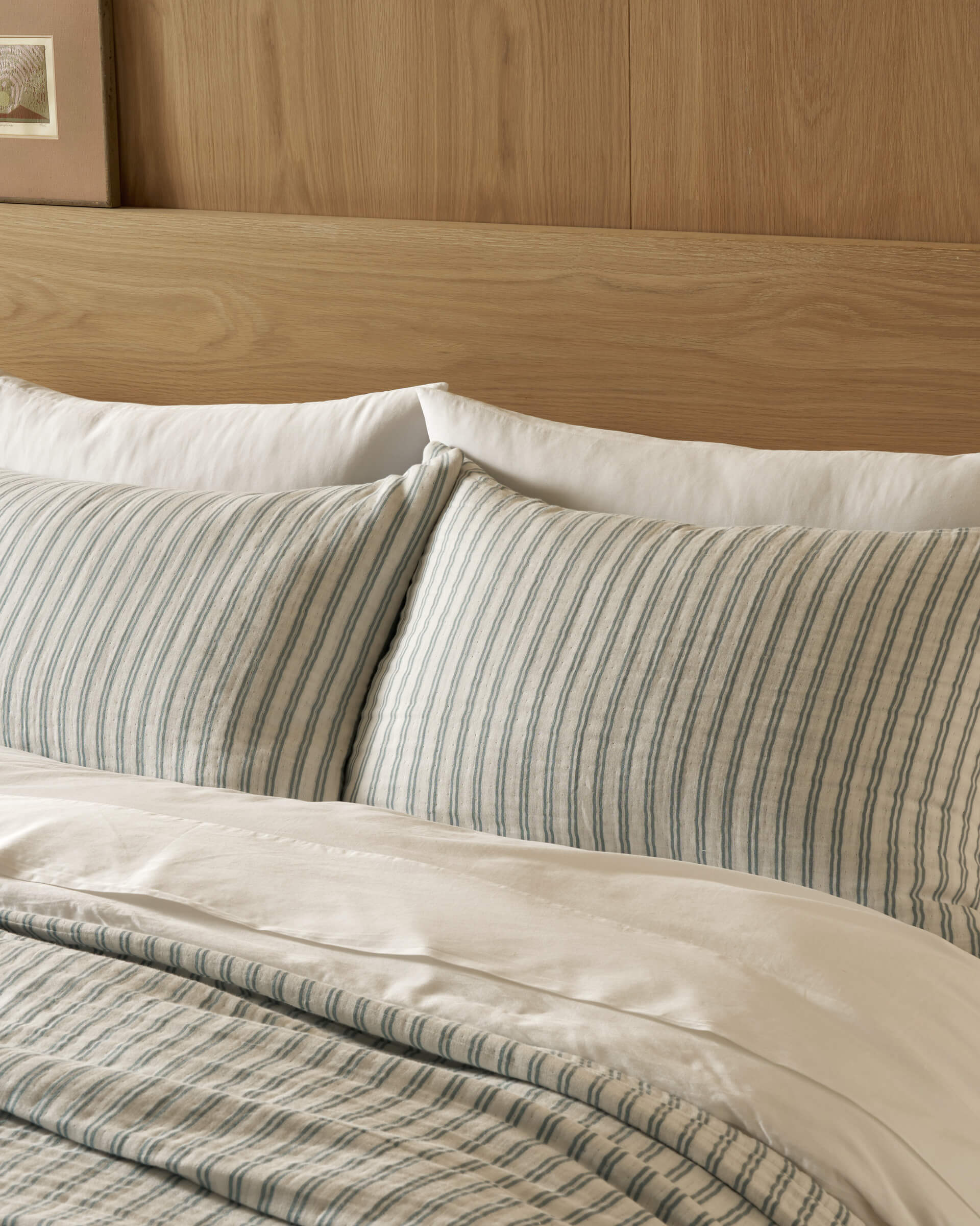 A bed made with Cozy Earth Waterfront Shams features white pillows and blue-and-white striped pillowcases, set against a wooden headboard with a framed picture partially visible on the wall.|Color:River Stripe