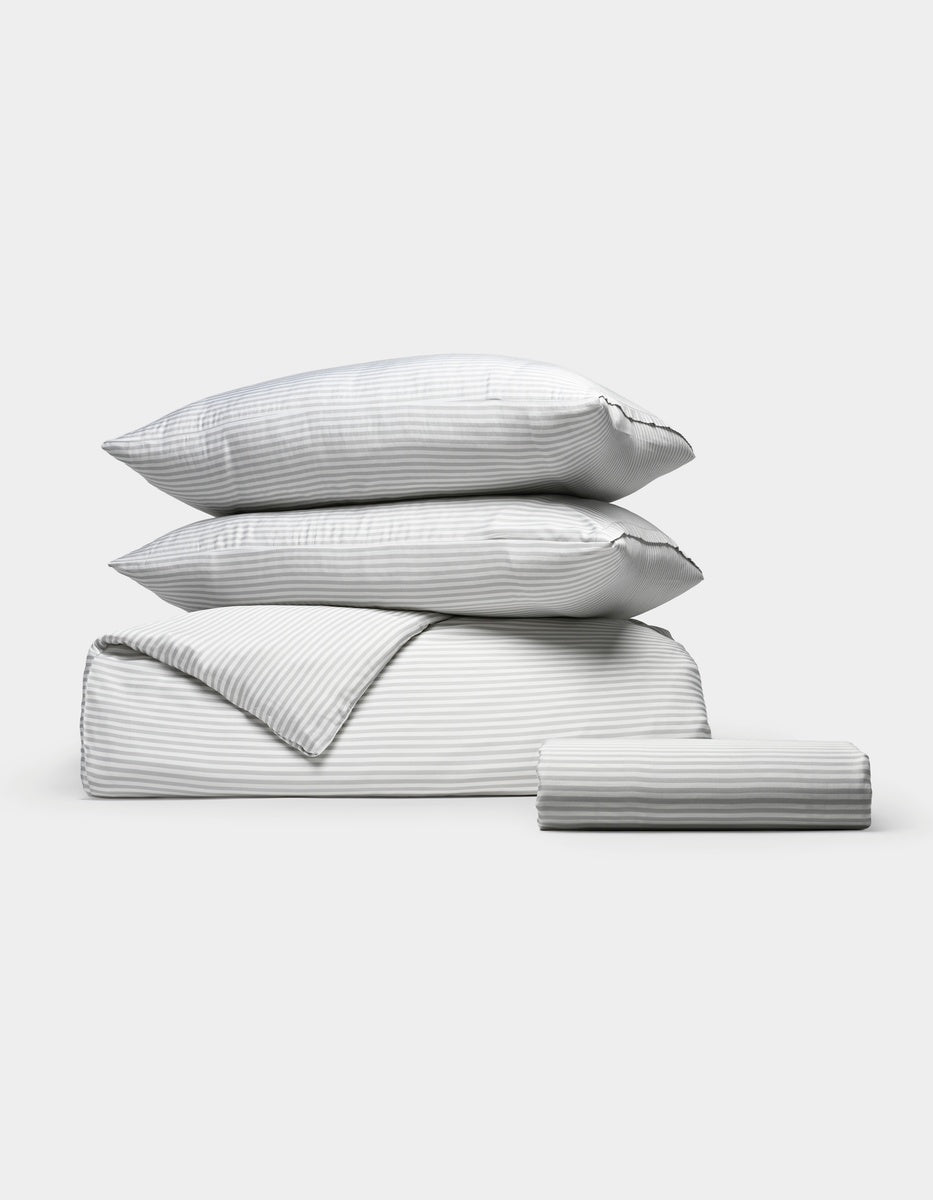 The Cozy Earth Bamboo Bedding Essential Bundle, featuring two stacked pillows, a folded comforter, and a folded sheet in light gray and white thin stripes, is arranged neatly against a plain white background.