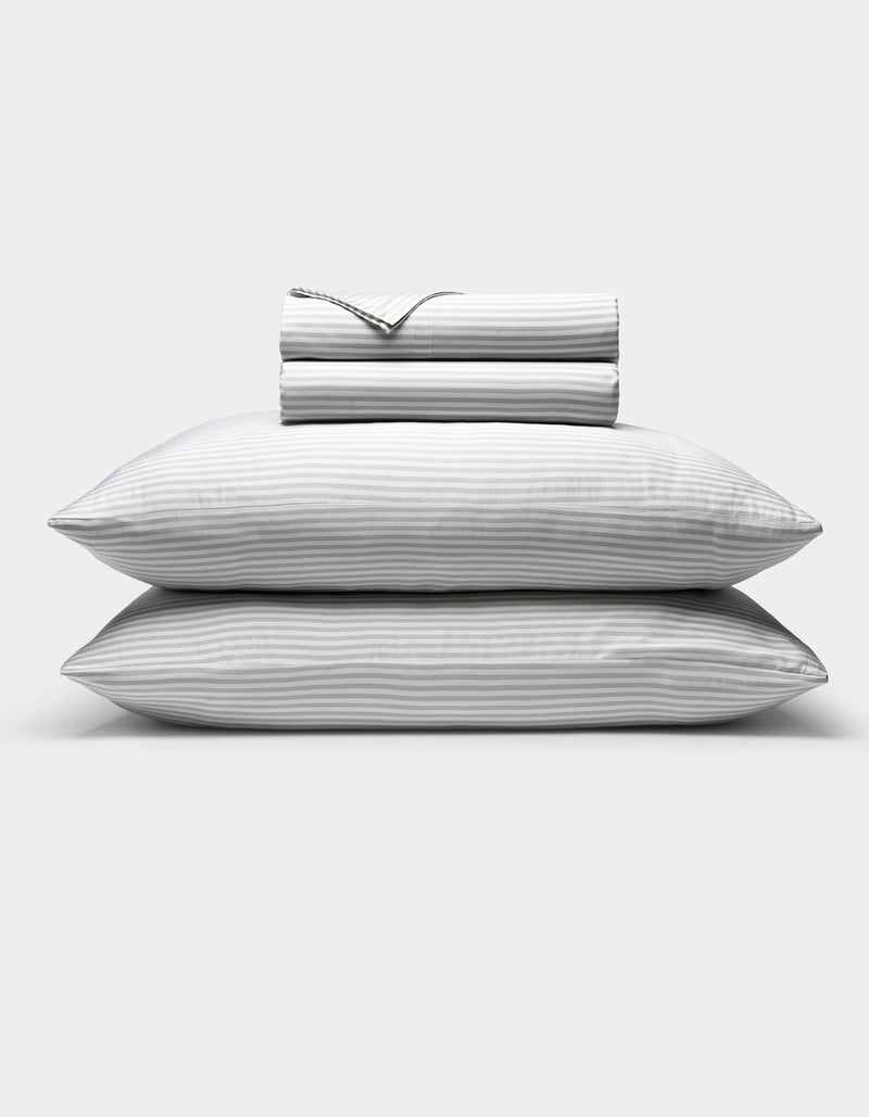 Two stacked pillows with subtle gray and white stripes are shown beneath a neatly folded Cozy Earth Bamboo Sheet Set, all displayed against a plain white background.