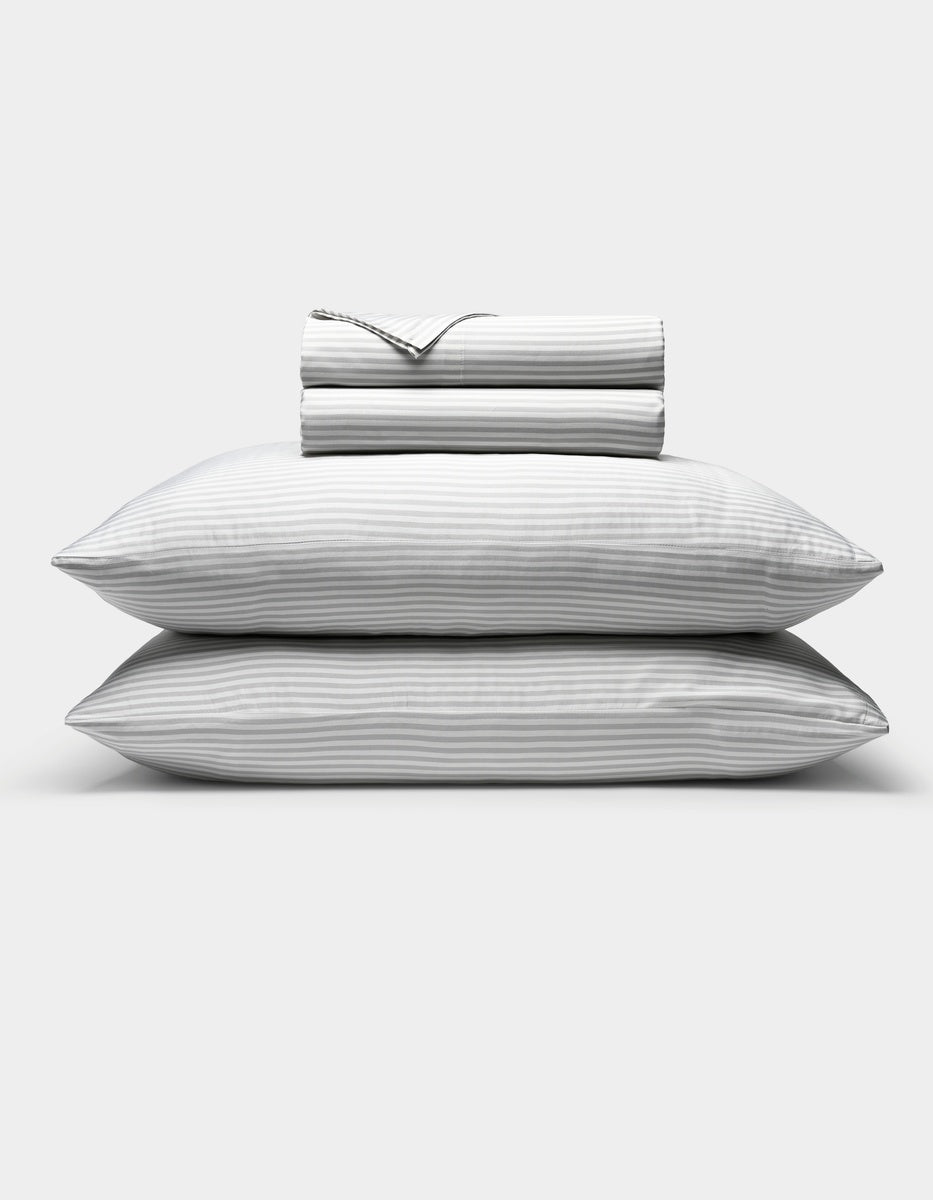 Two stacked pillows with subtle gray and white stripes are shown beneath a neatly folded Cozy Earth Bamboo Sheet Set, all displayed against a plain white background.|Color:Classic Stripe in Dew