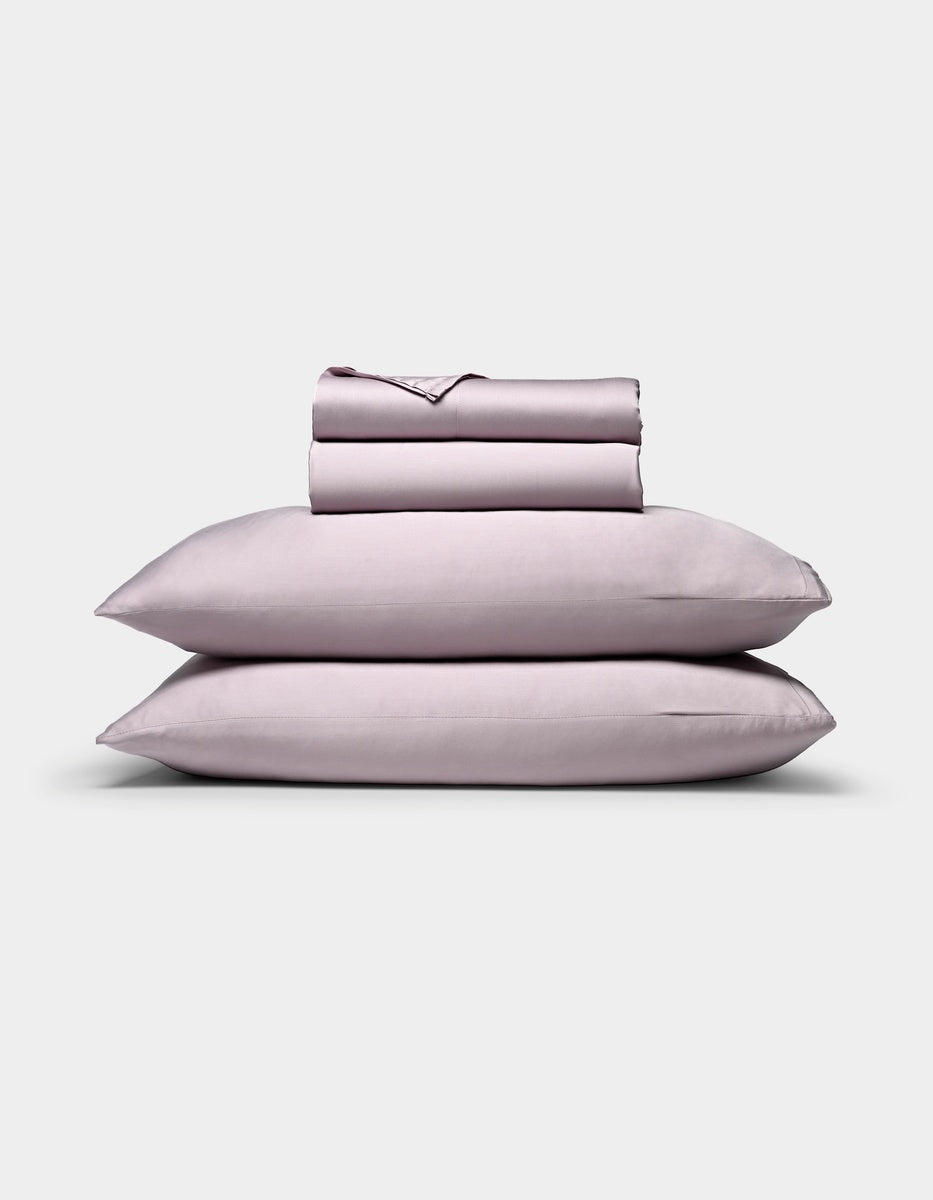 A neatly folded Bamboo Sheet Set by Cozy Earth in light lavender, featuring two stacked pillows, a fitted sheet, a flat sheet, and pillowcases, displayed against a plain white background.|Color:Lavender
