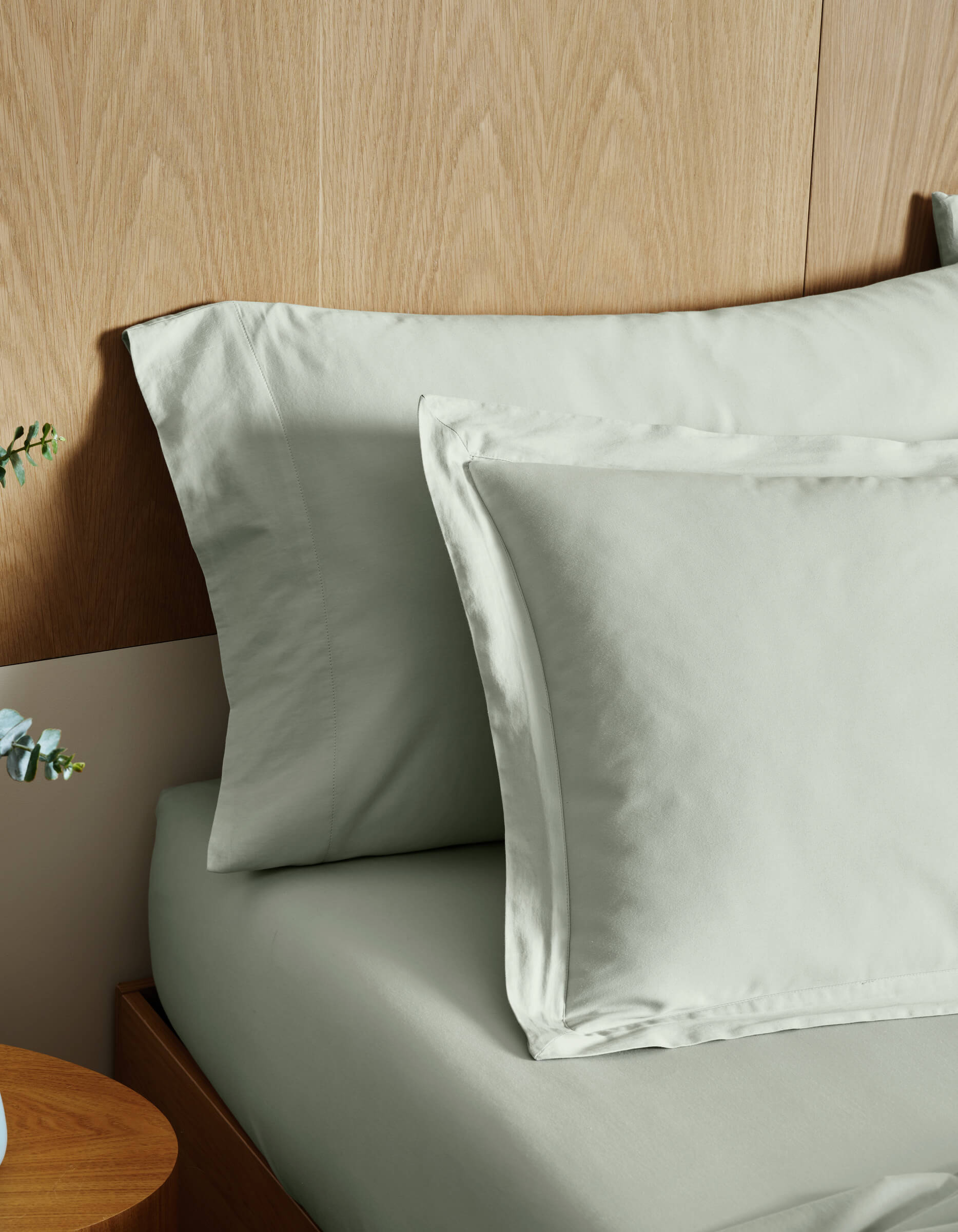 Cozy Earth Soft-Wash Cotton Shams in light gray are neatly arranged on a wooden bed with a wood-paneled headboard; part of a wooden nightstand and a small plant appear in the foreground.|Color:Laurel