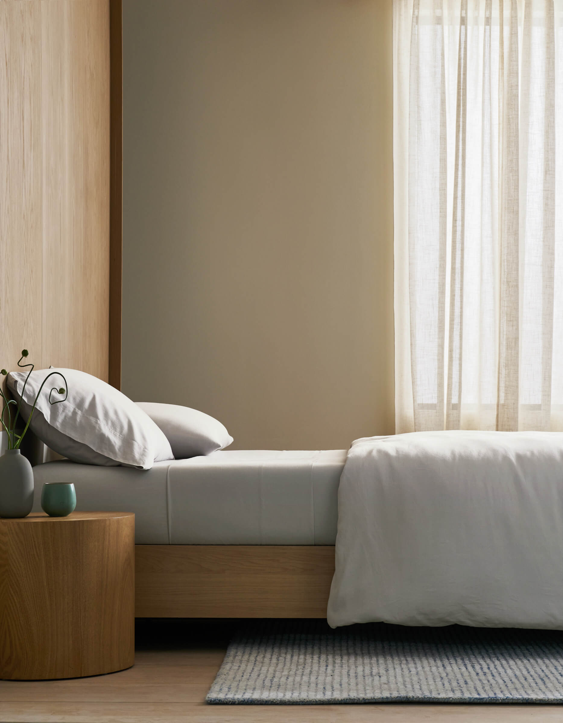 A minimalist bedroom features a neatly made bed with Cozy Earth's Soft-Wash Cotton Bedding Core Bundle, white bedding, and pillows beside a wooden nightstand with a vase and cup. Sheer curtains filter soft natural light, creating a serene ambiance.|Color:Dew