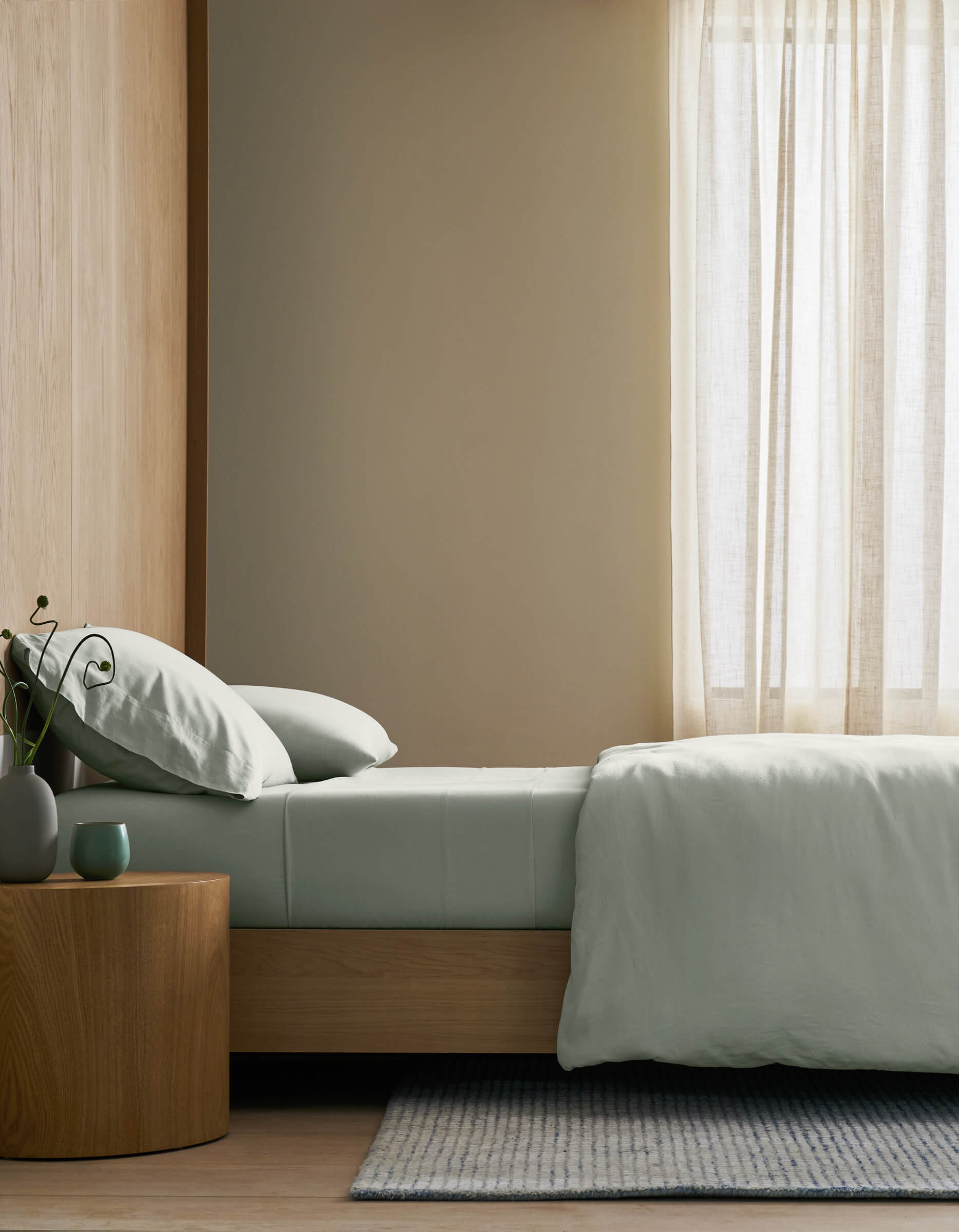 A minimalist bedroom featuring a neatly made bed with Cozy Earth's Soft-Wash Cotton Bedding Core Bundle in pale green, a round wooden nightstand with a plant and cup, light wood floors, and sheer curtains letting in gentle natural light.|Color:Laurel