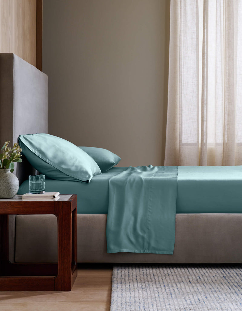A neatly made bed with Cozy Earth's Bamboo Fitted Sheet in light teal and matching pillows sits beside a wooden nightstand holding a glass of water and flowers, all in a softly lit, modern bedroom with beige walls and curtains.