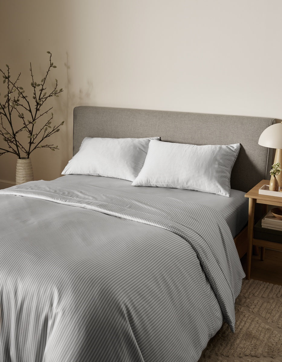 A neatly made bed featuring the Bamboo Bedding Core Bundle from Cozy Earth in light gray stripes, topped with two pillows and set against a gray upholstered headboard, is complemented by a potted branch, lamp, and wooden nightstand in a cozy, minimalistic bedroom.