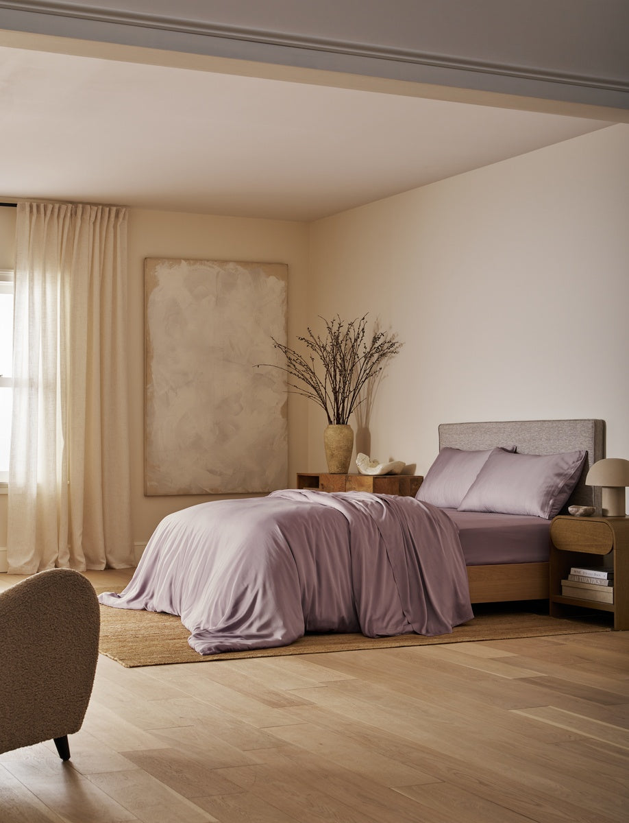 A bedroom scene with a bed dressed in soft lavender bedding, set in a warm neutral room with beige walls, wood flooring, curtains, and a large abstract painting.