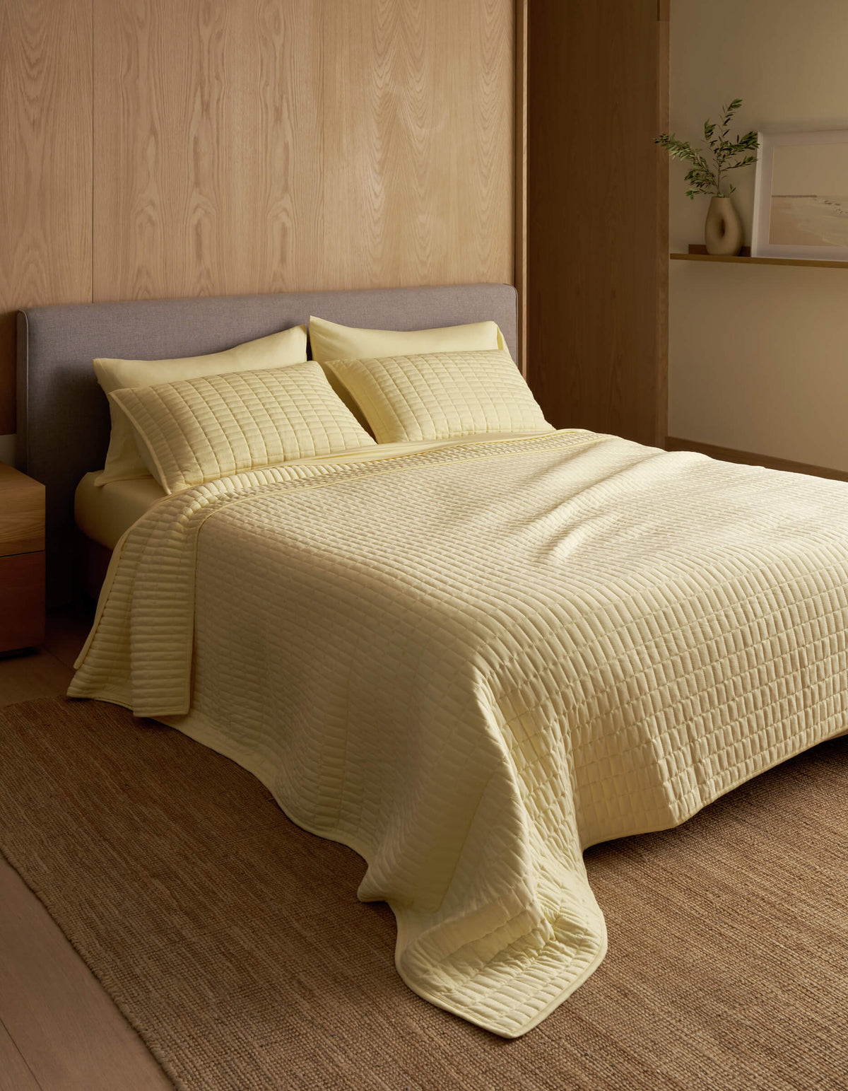 A minimalist bedroom features a neatly made bed with the Cozy Earth Bamboo Jersey Quilt in pale yellow, matching pillows, wood-paneled walls, a neutral rug, and a small shelf displaying a vase and framed art.|Color:Meringue