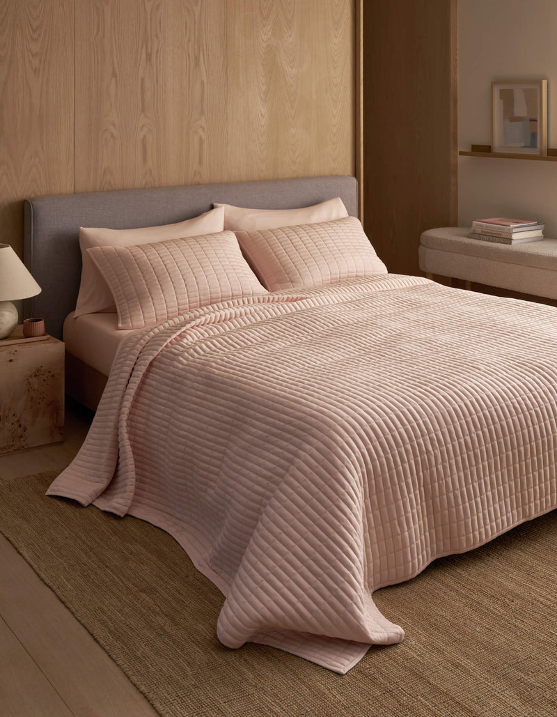 A neatly made bed features the Cozy Earth Bamboo Jersey Quilt in light pink with matching pillows, set against a wooden wall in a minimal, modern bedroom with neutral tones and simple decor.