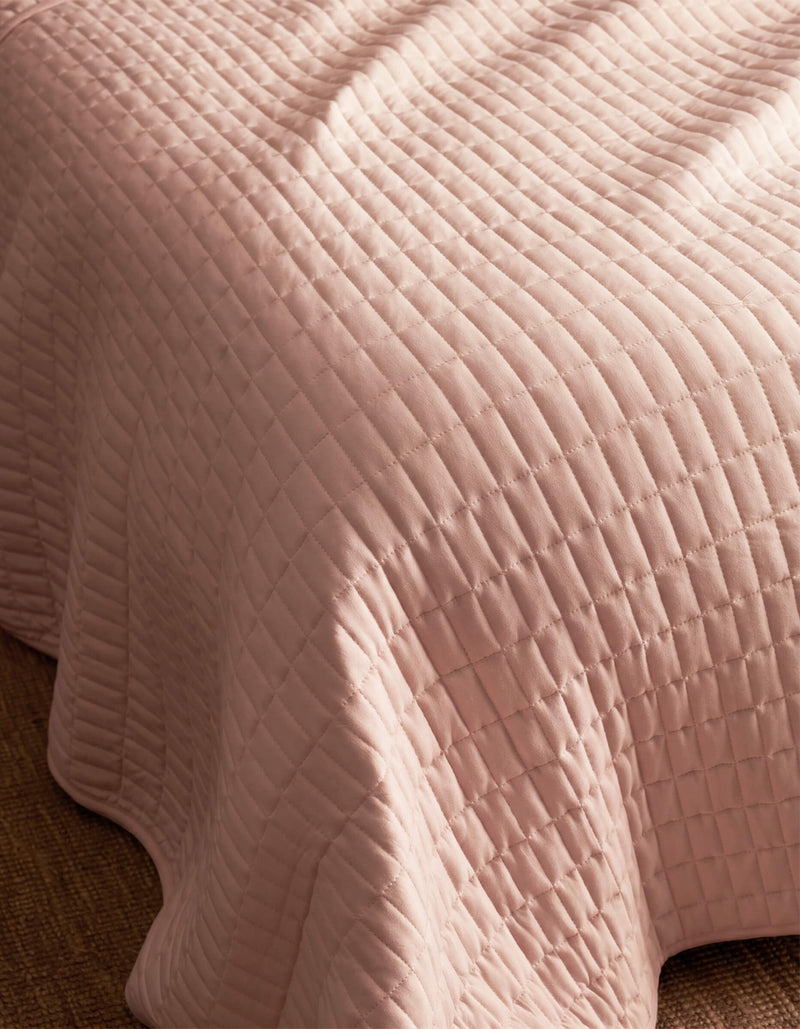 A close-up of the Cozy Earth Bamboo Jersey Quilt Bundle in pale pink with a quilted grid pattern, draped over a bed’s edge and resting on a textured brown carpet.