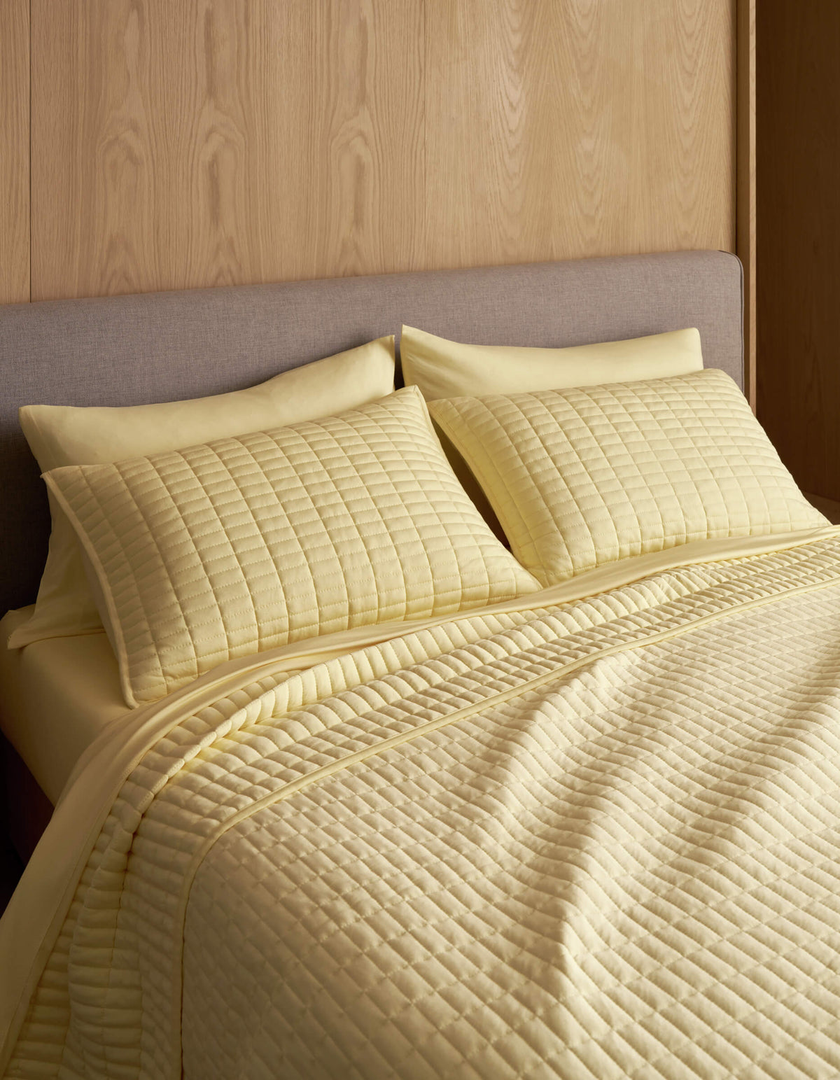 A neatly made bed features the Cozy Earth Bamboo Jersey Quilt in light yellow with matching pillow shams, set against a light wood paneled wall and a gray upholstered headboard.|Color:Meringue