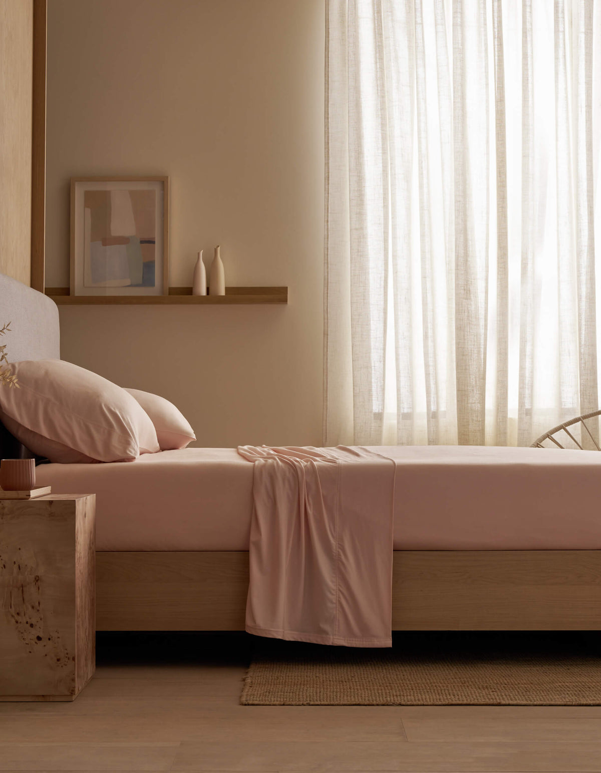 A minimal bedroom features a neatly made bed with Cozy Earth's Bamboo Jersey Sheet Set in pale pink, soft light filtered through sheer curtains, a wooden side table, framed abstract art, and two vases on a wall shelf.|Color:Petal
